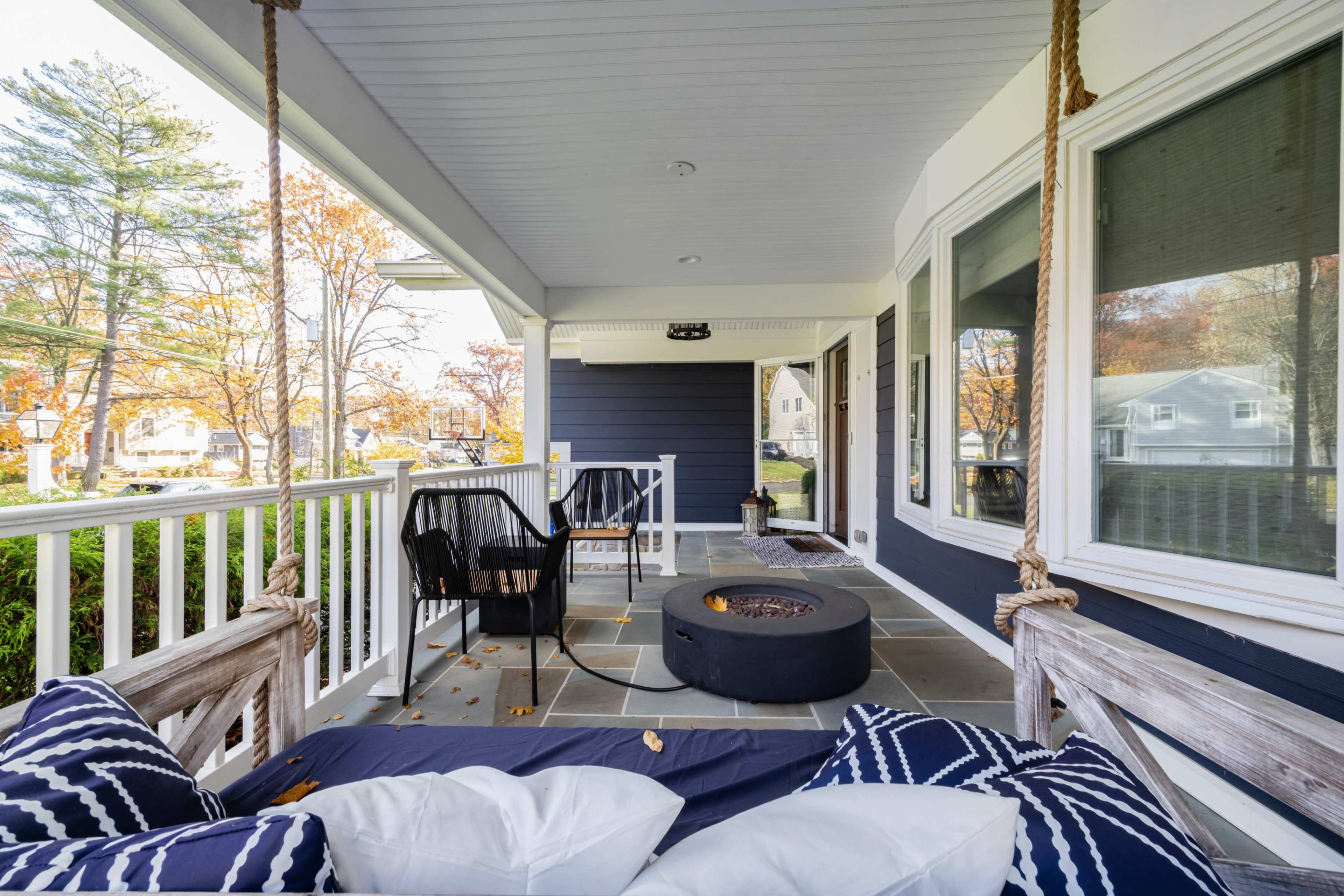 The image shows a porch with a swing bench, two black chairs, and a circular table, surrounded by greenery and autumn trees.