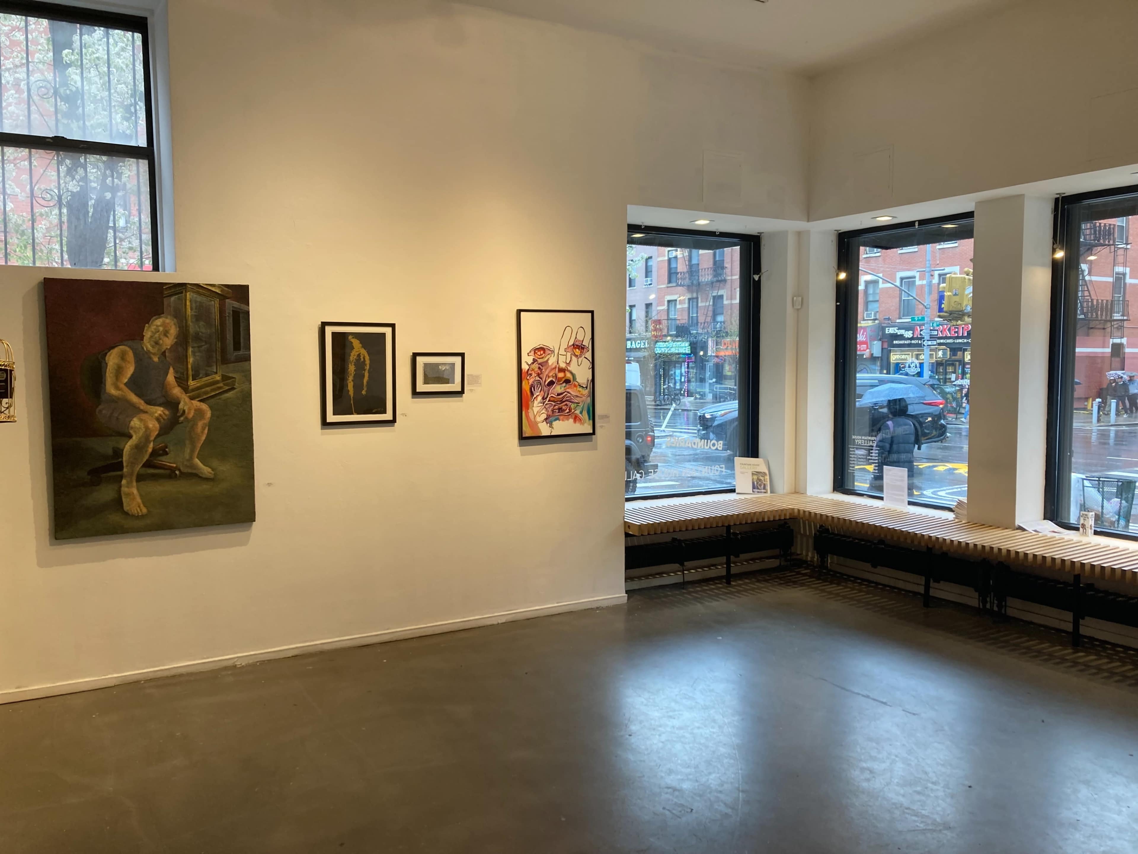 The image shows an art gallery interior with several framed artworks displayed on white walls and large windows revealing a street view.