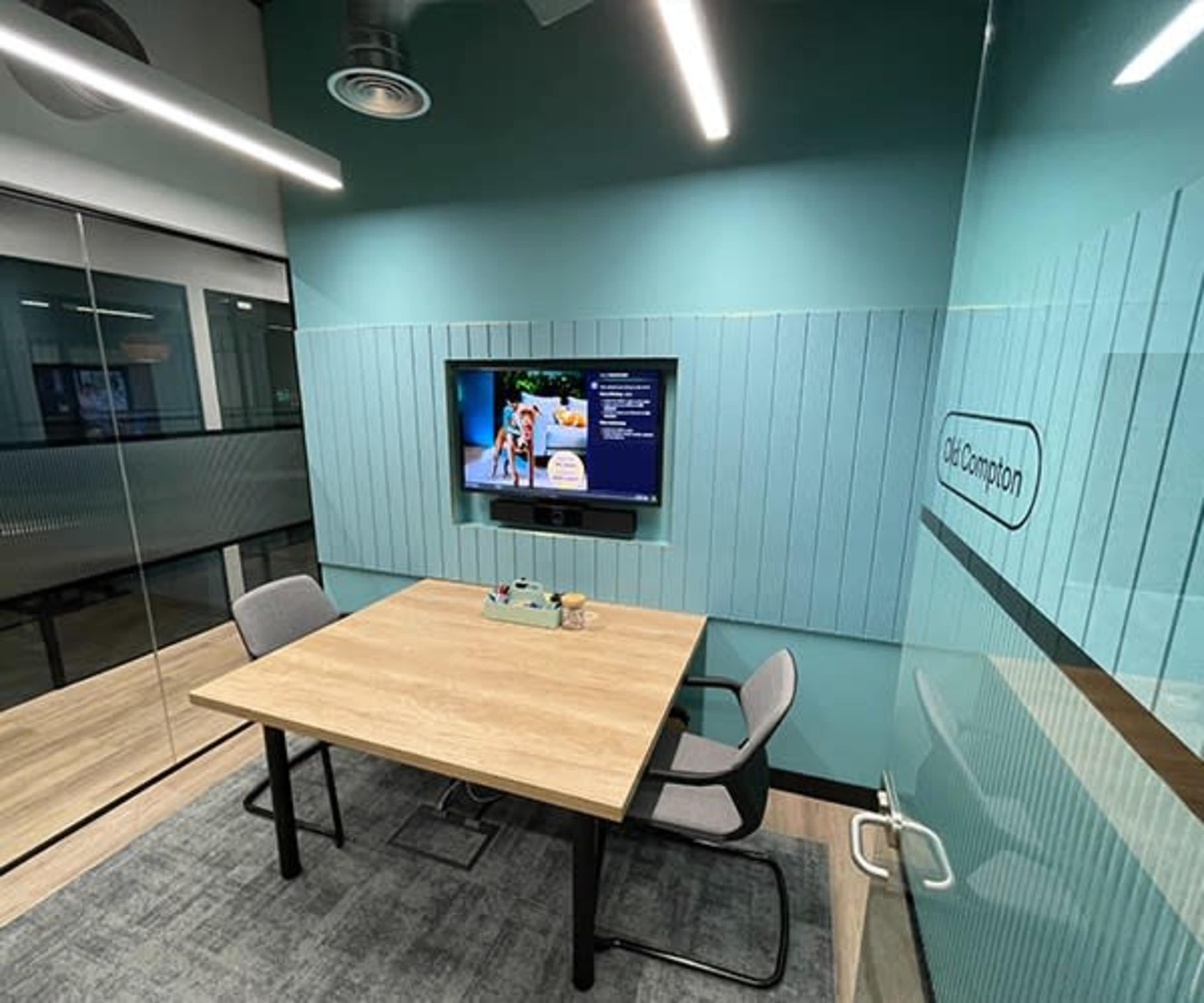 An office meeting room features a wooden table with two chairs, a television mounted on a wall, and glass partitions.