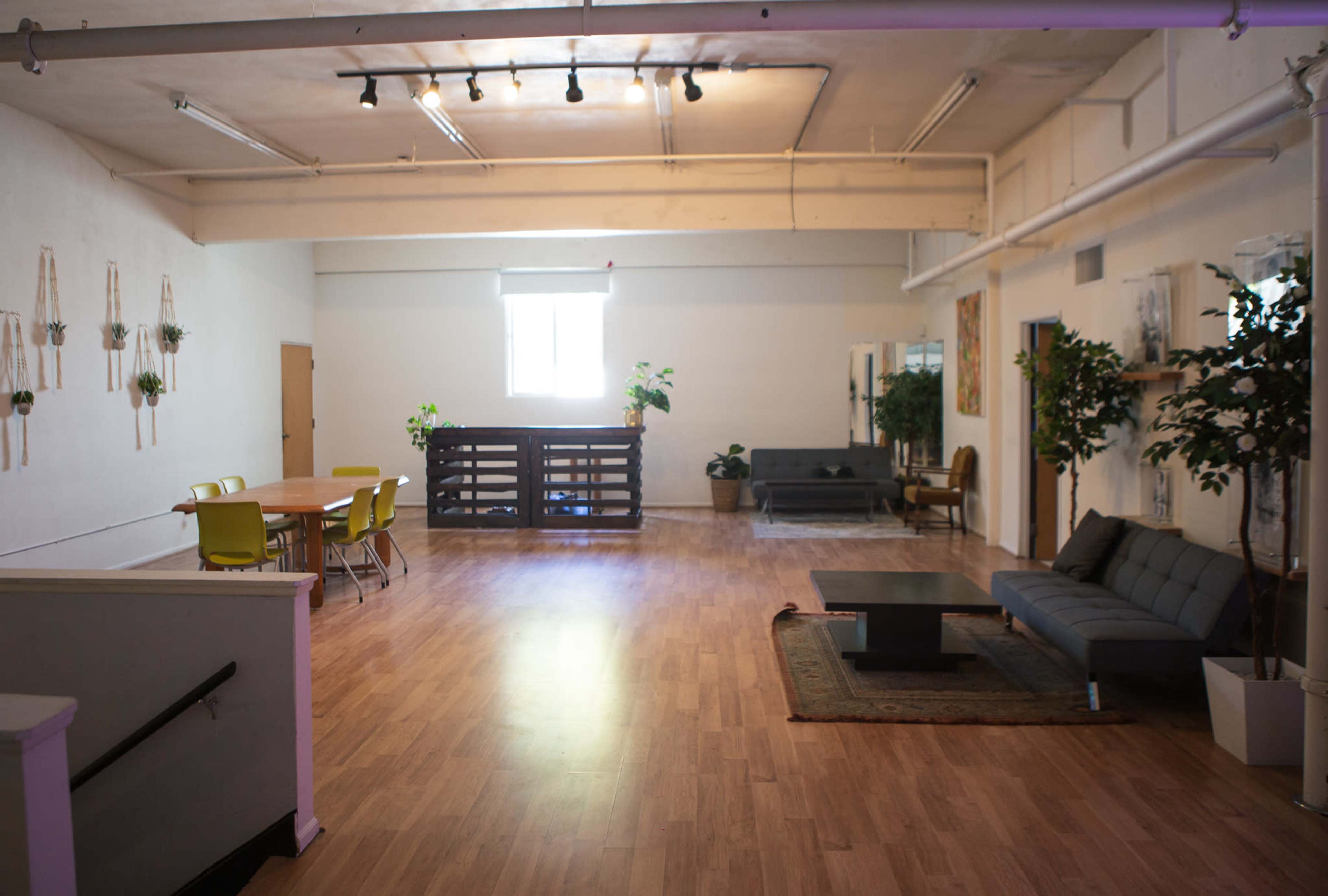 A spacious interior with light wood flooring, featuring a dining area with yellow chairs, a seating area with a gray couch, and several plants placed around the room.