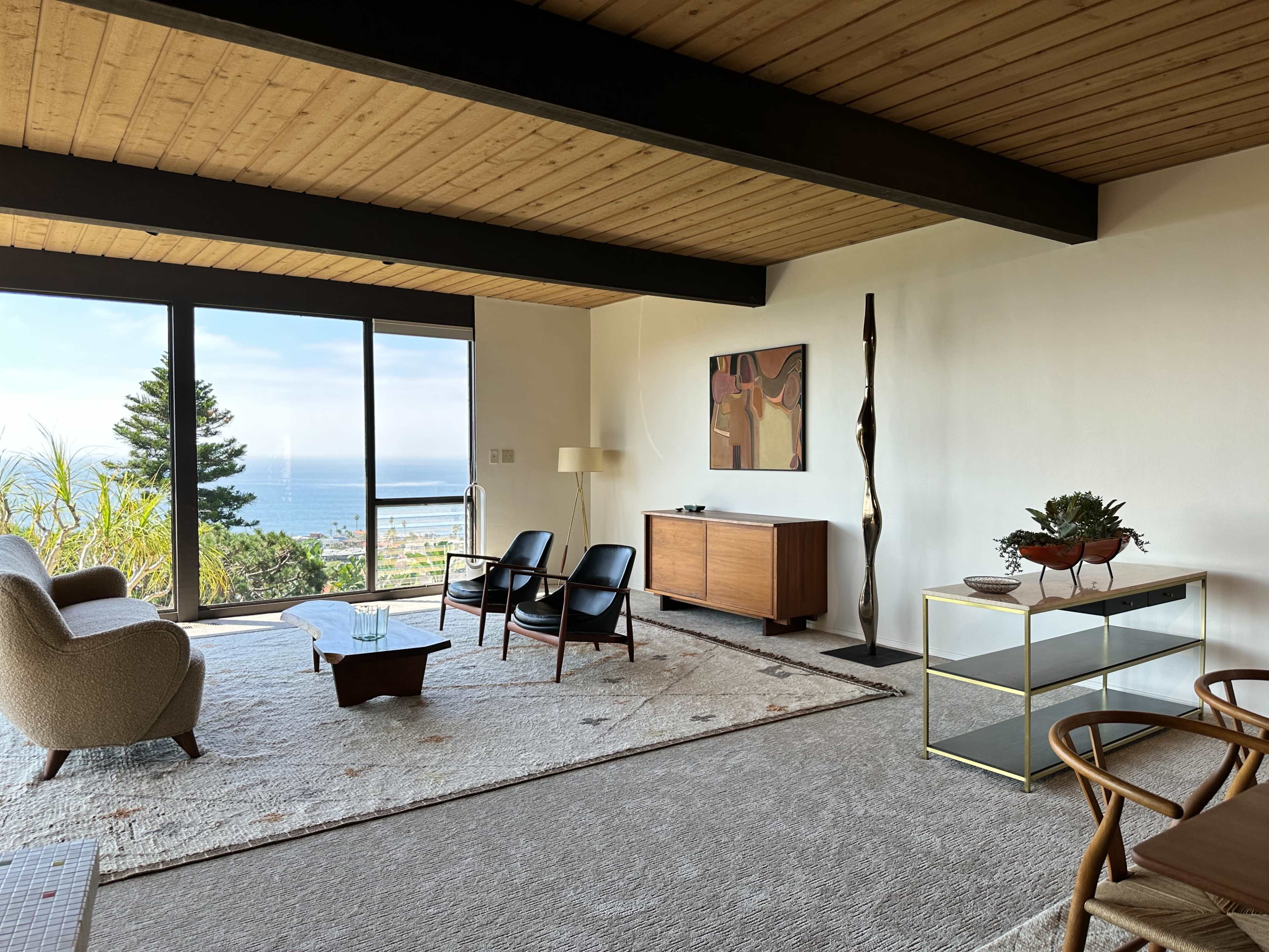 A modern living room features a large window with an ocean view, two black chairs, a beige sofa, a wooden sideboard, and a decorative plant on a glass table.