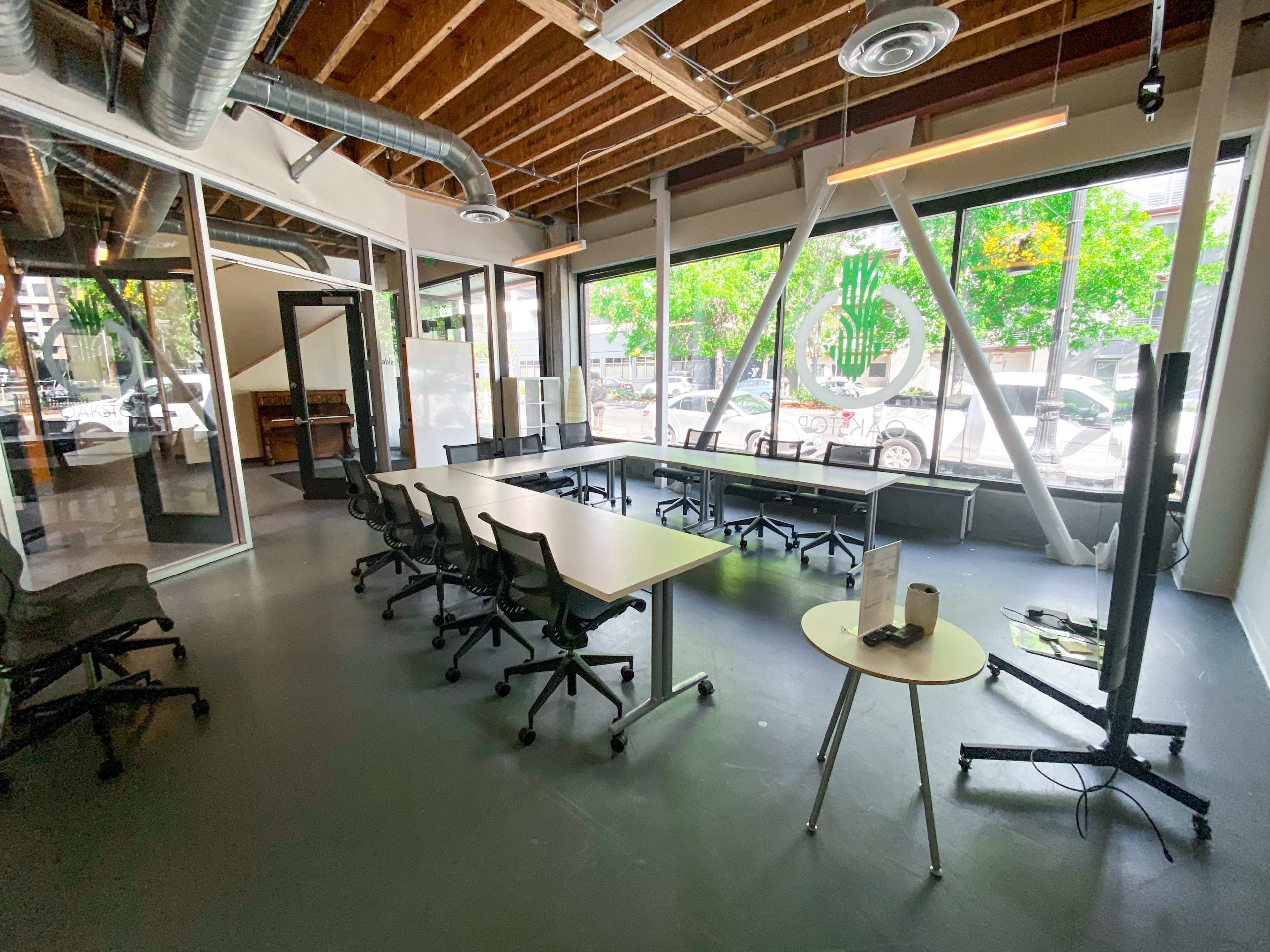 A modern office workspace features several tables and chairs arranged in a bright area with large windows and exposed beams.