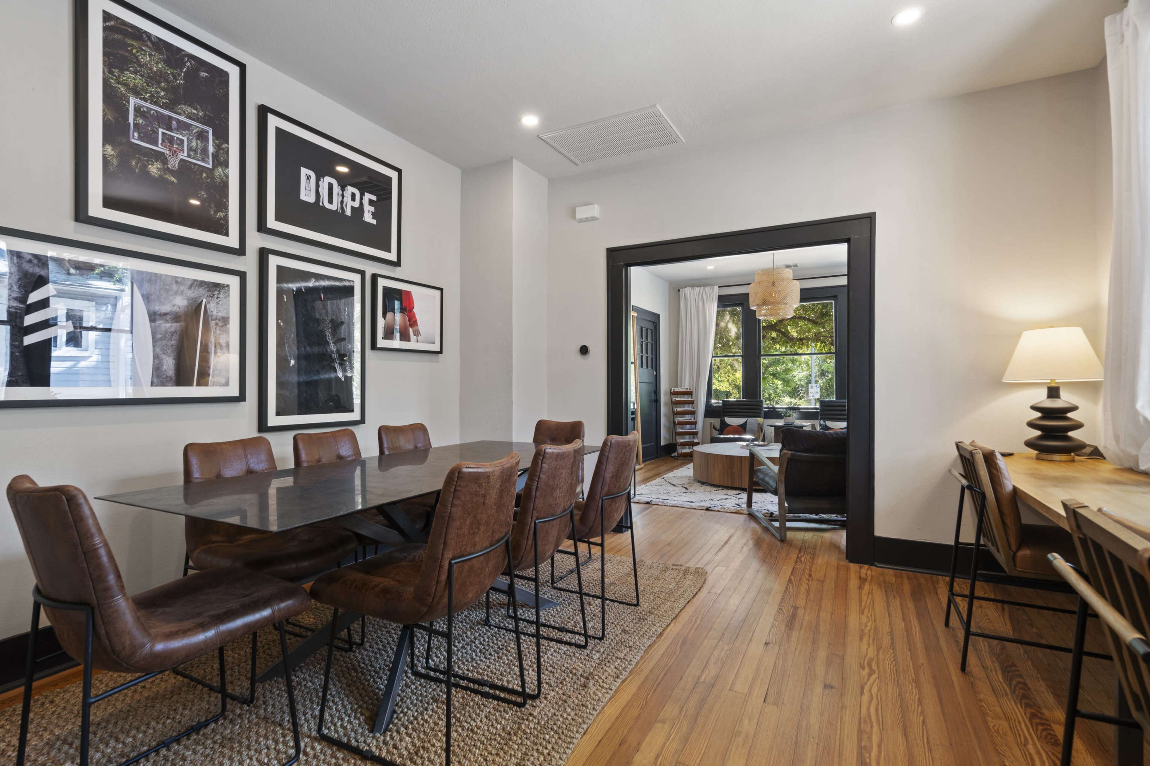 A dining area with a large table surrounded by brown chairs, framed artwork on the walls, and a view into a living room with large windows.