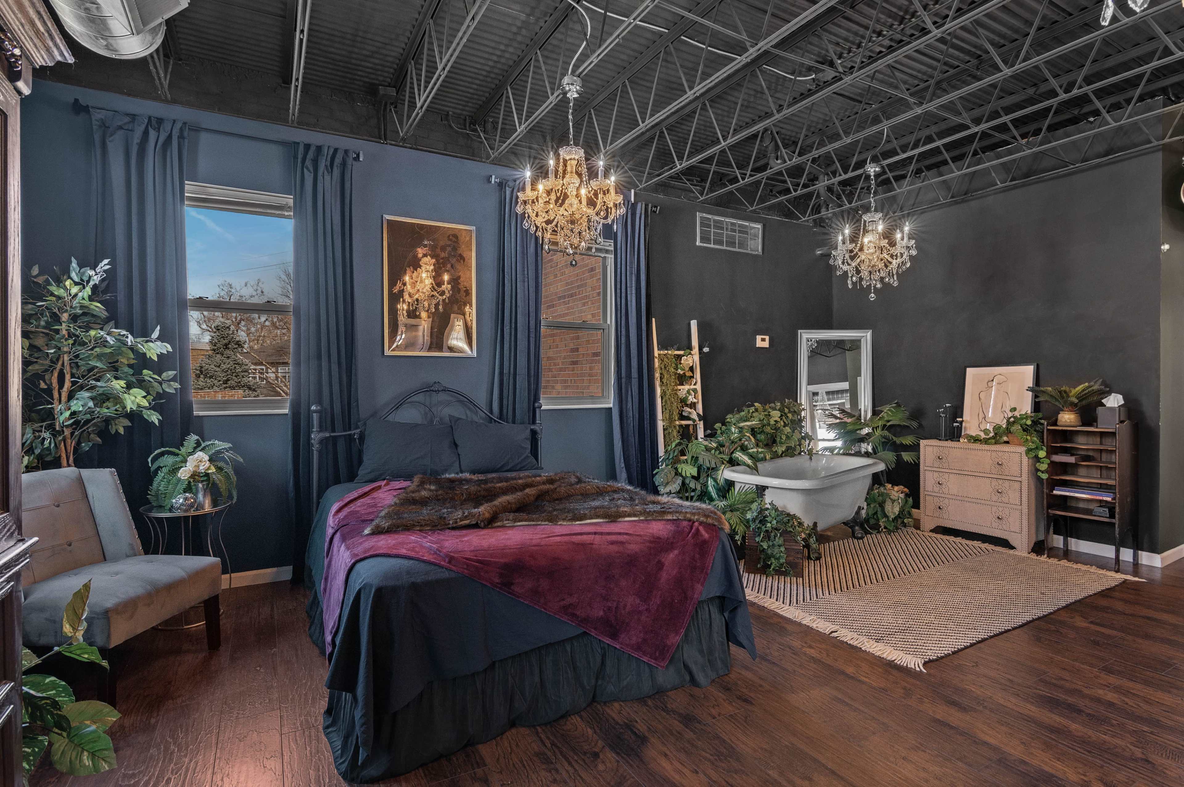 A dimly lit bedroom featuring a bed with a dark cover, a freestanding bathtub, and various plants arranged throughout the space.