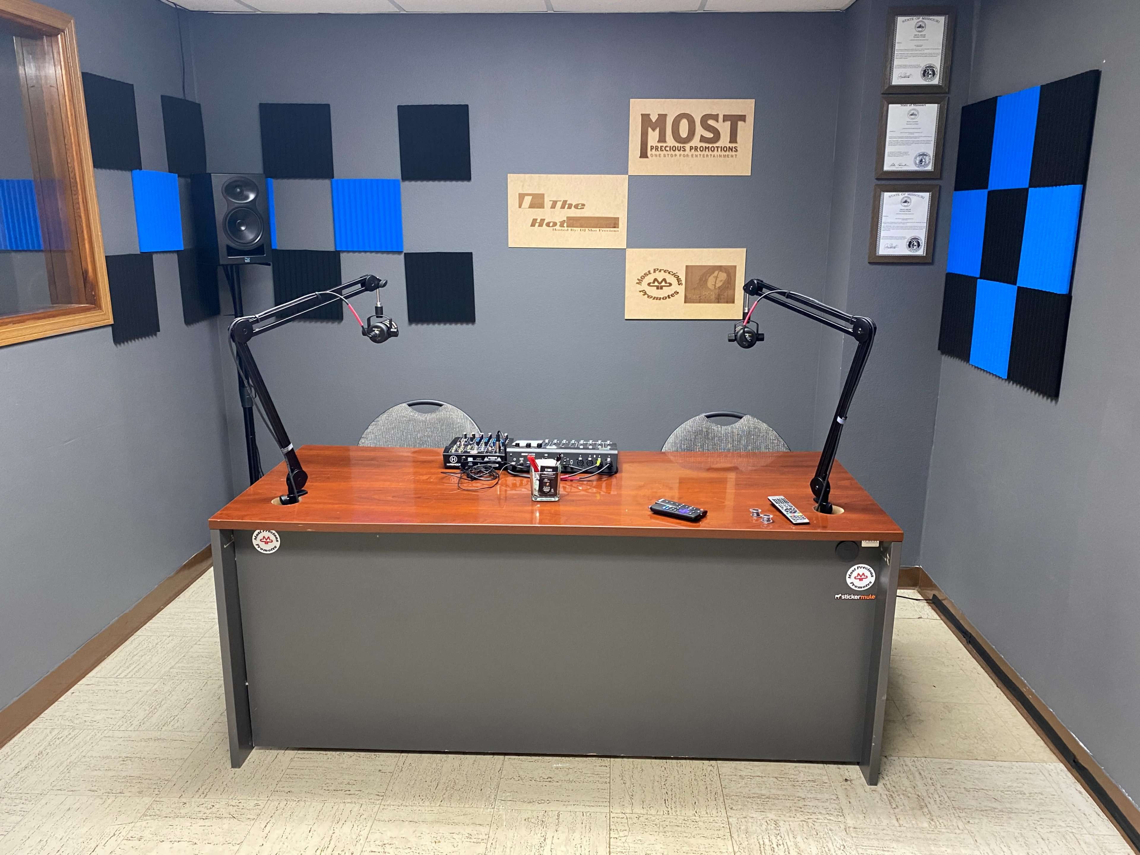 A recording studio with a wooden table, two microphones on adjustable arms, a mixing console, and soundproofing panels on the walls.