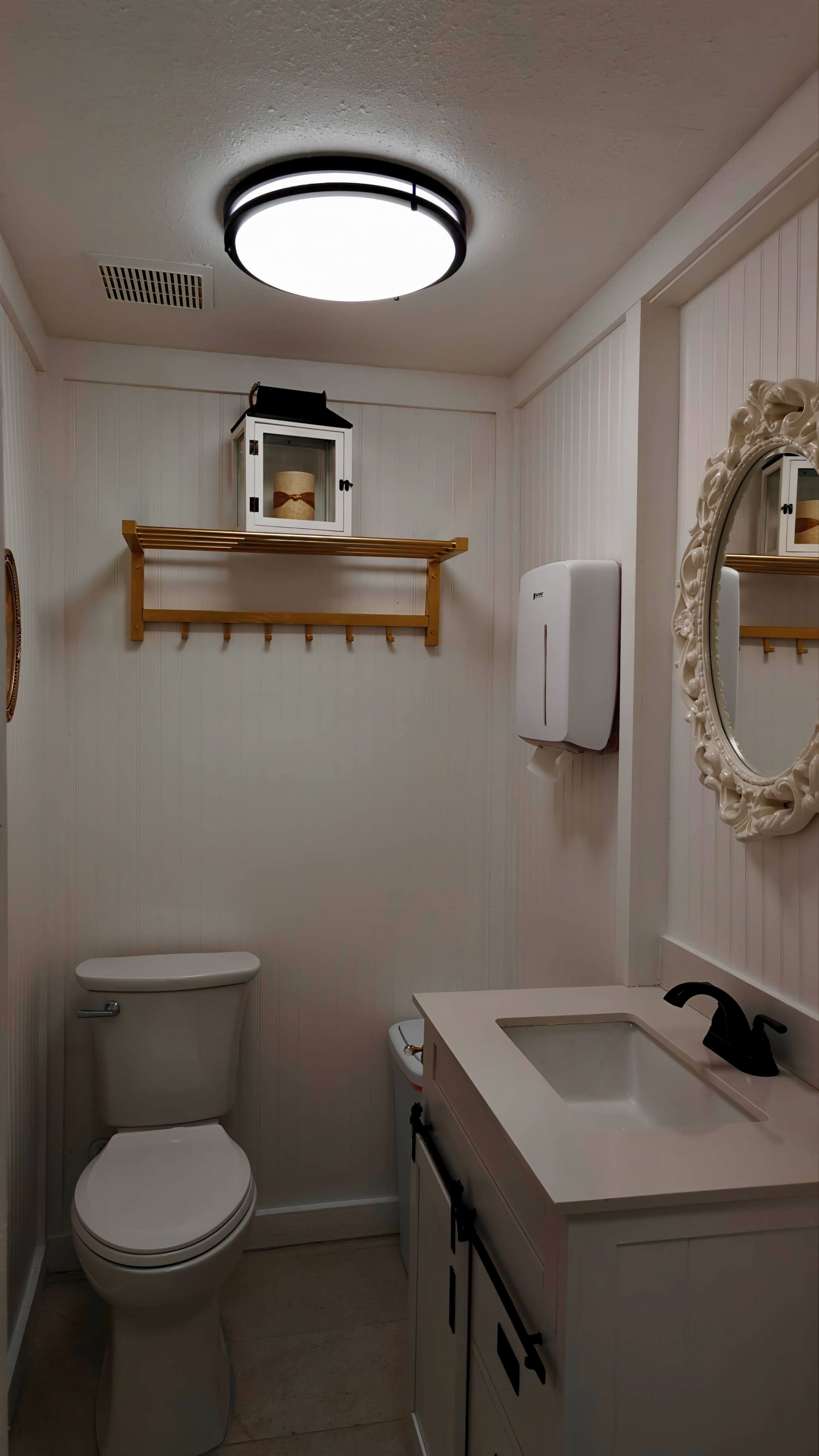 The image shows a small bathroom with a toilet, a white sink with a black faucet, a wall-mounted soap dispenser, a round mirror, and a shelf holding a decorative lantern.