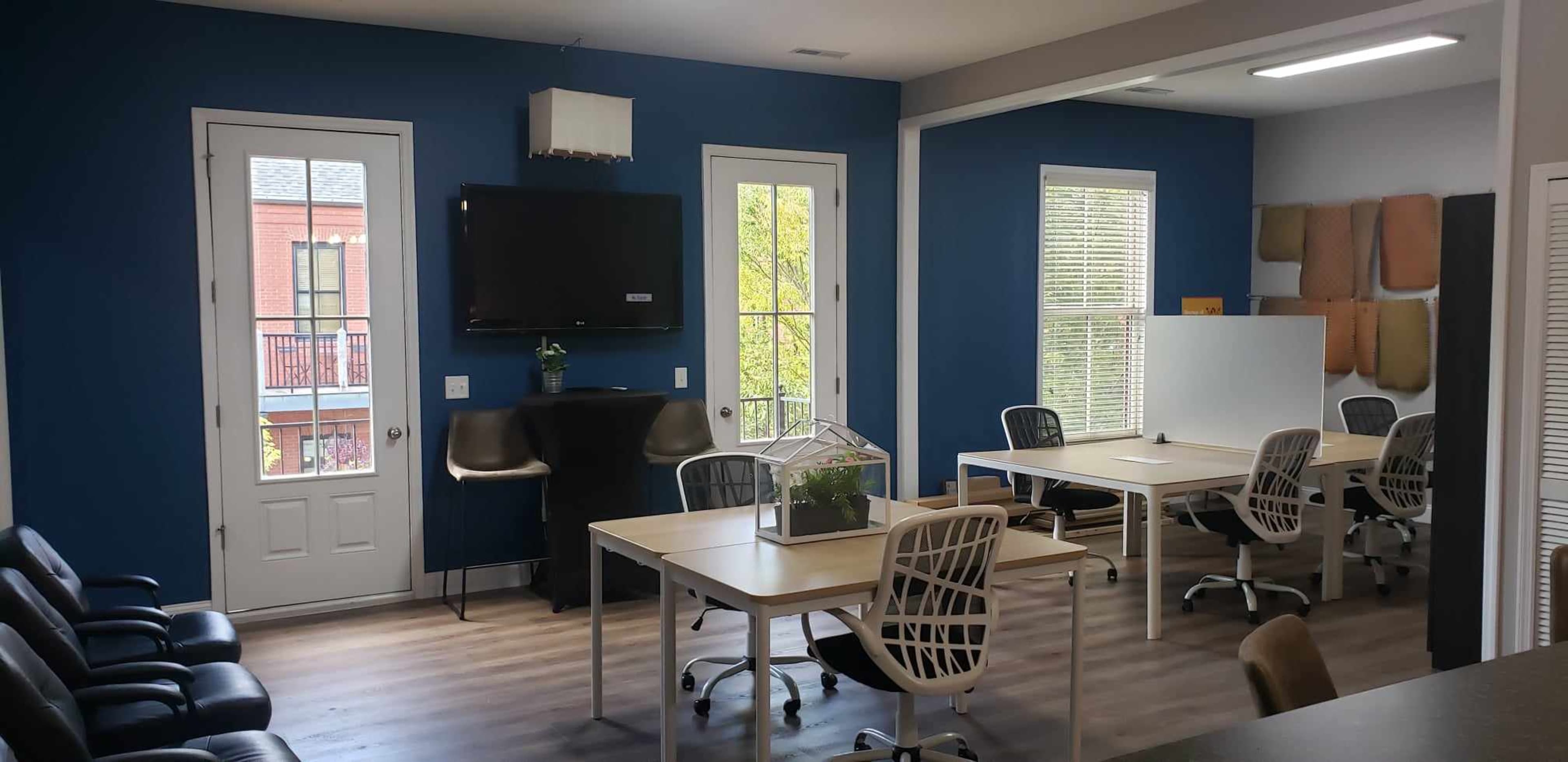 A room with blue walls features several tables and chairs, a television on the wall, and large windows providing natural light.