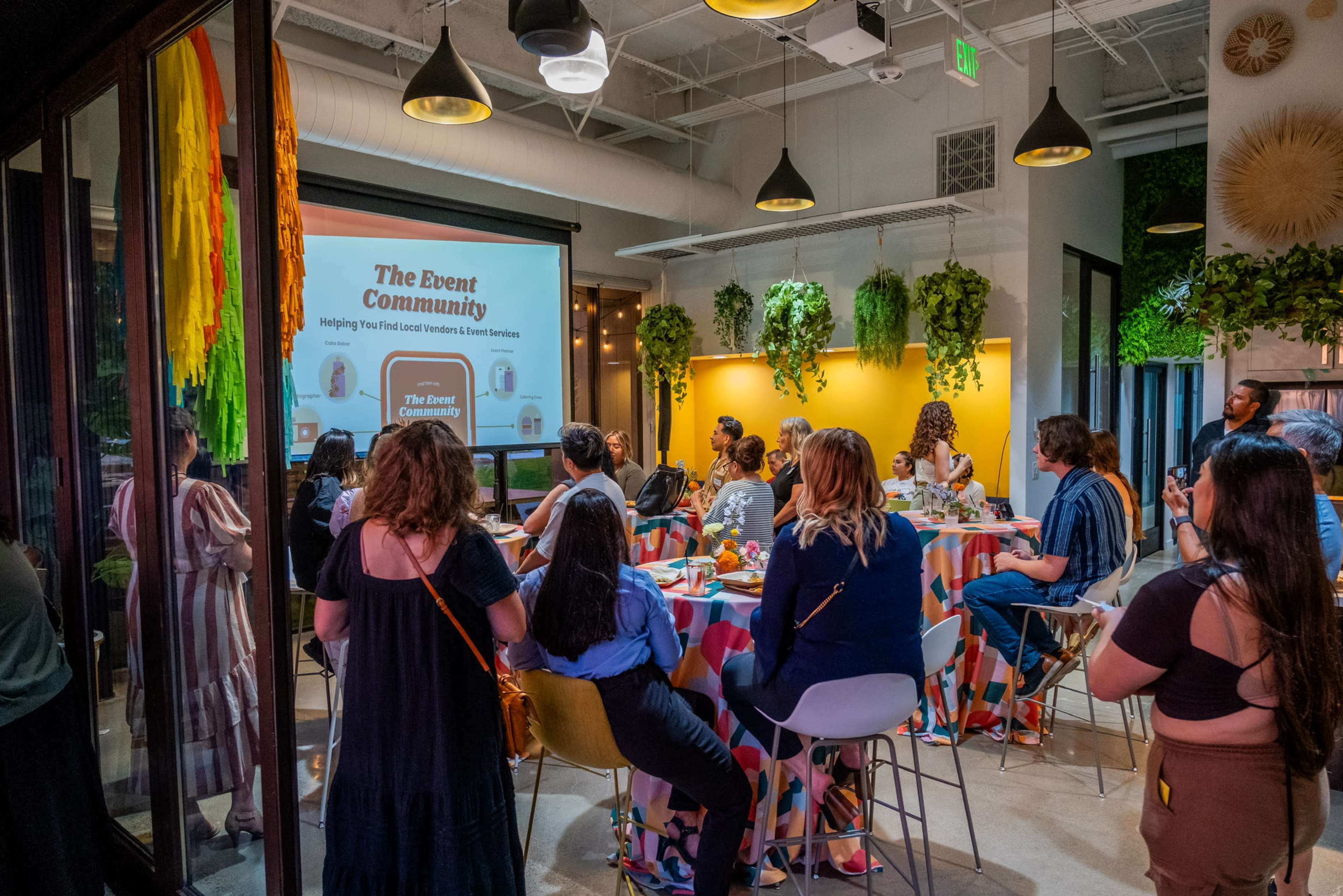 A group of people is seated in a colorful space, attentively watching a presentation on a large screen during an event.