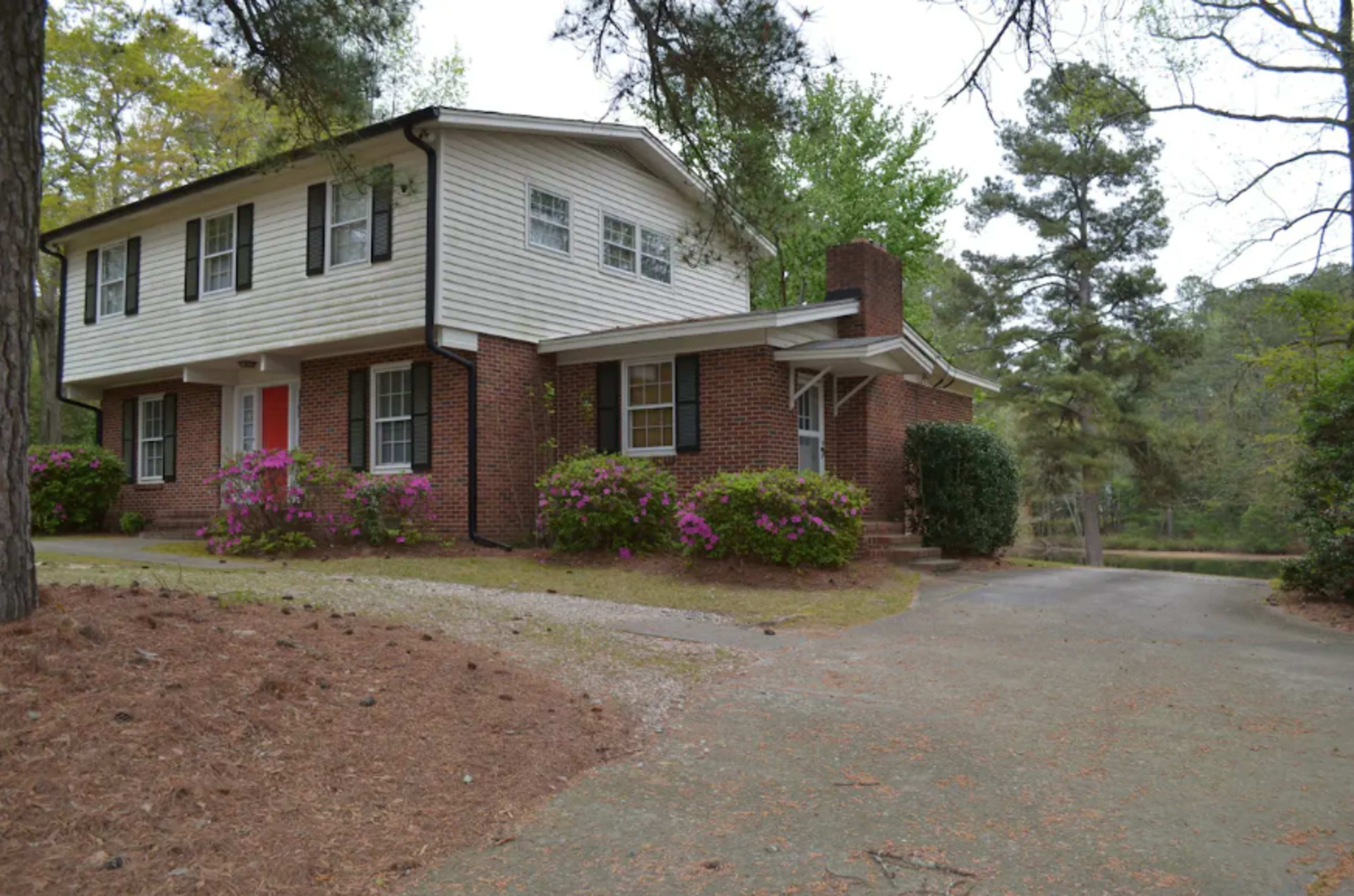 The image shows a two-story house with white siding and a brick foundation, surrounded by green trees and flowering bushes along a curved driveway.