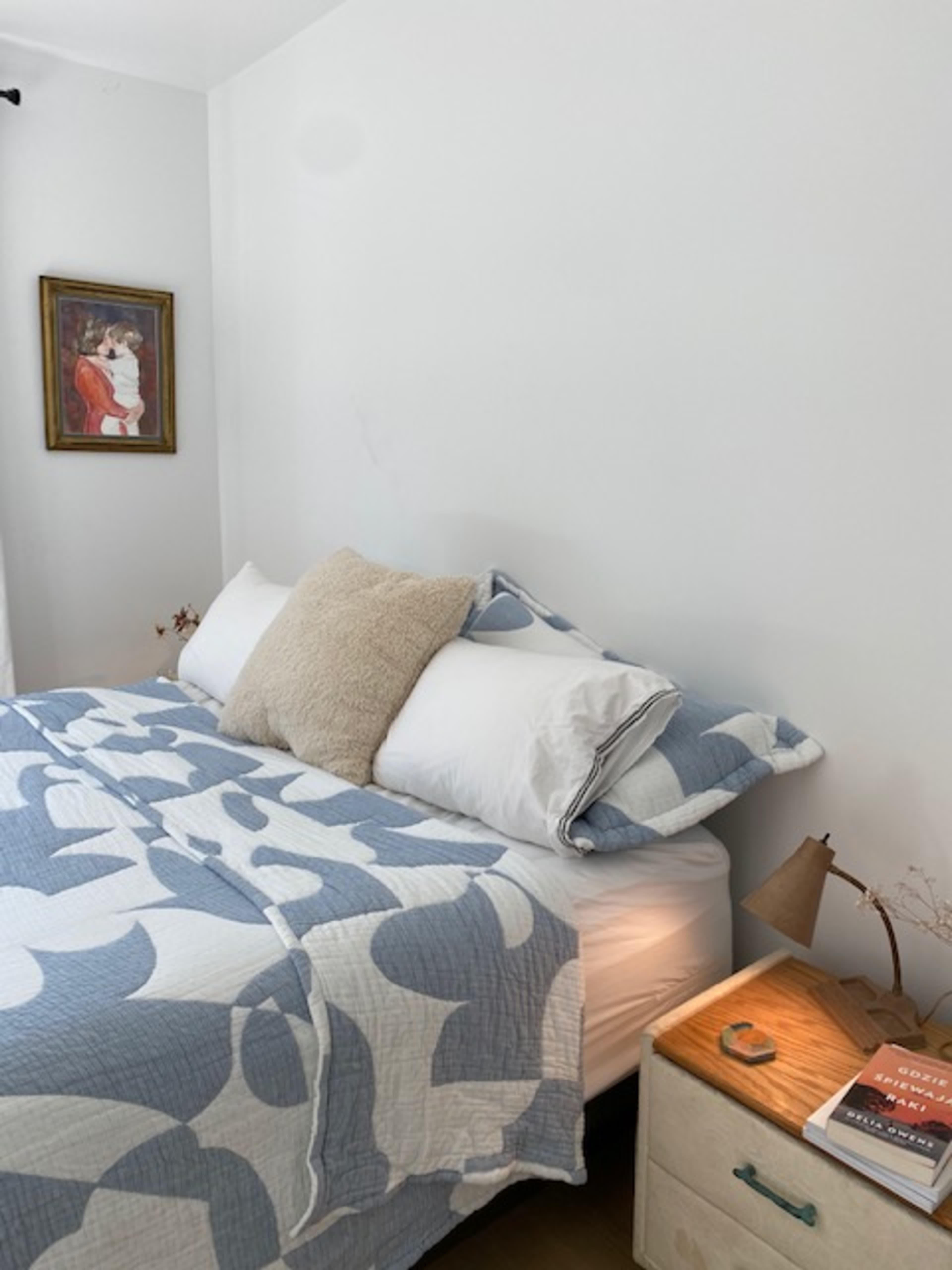 A bed with a blue and white patterned quilt is positioned against a white wall, accompanied by a bedside table holding a lamp and a few books.