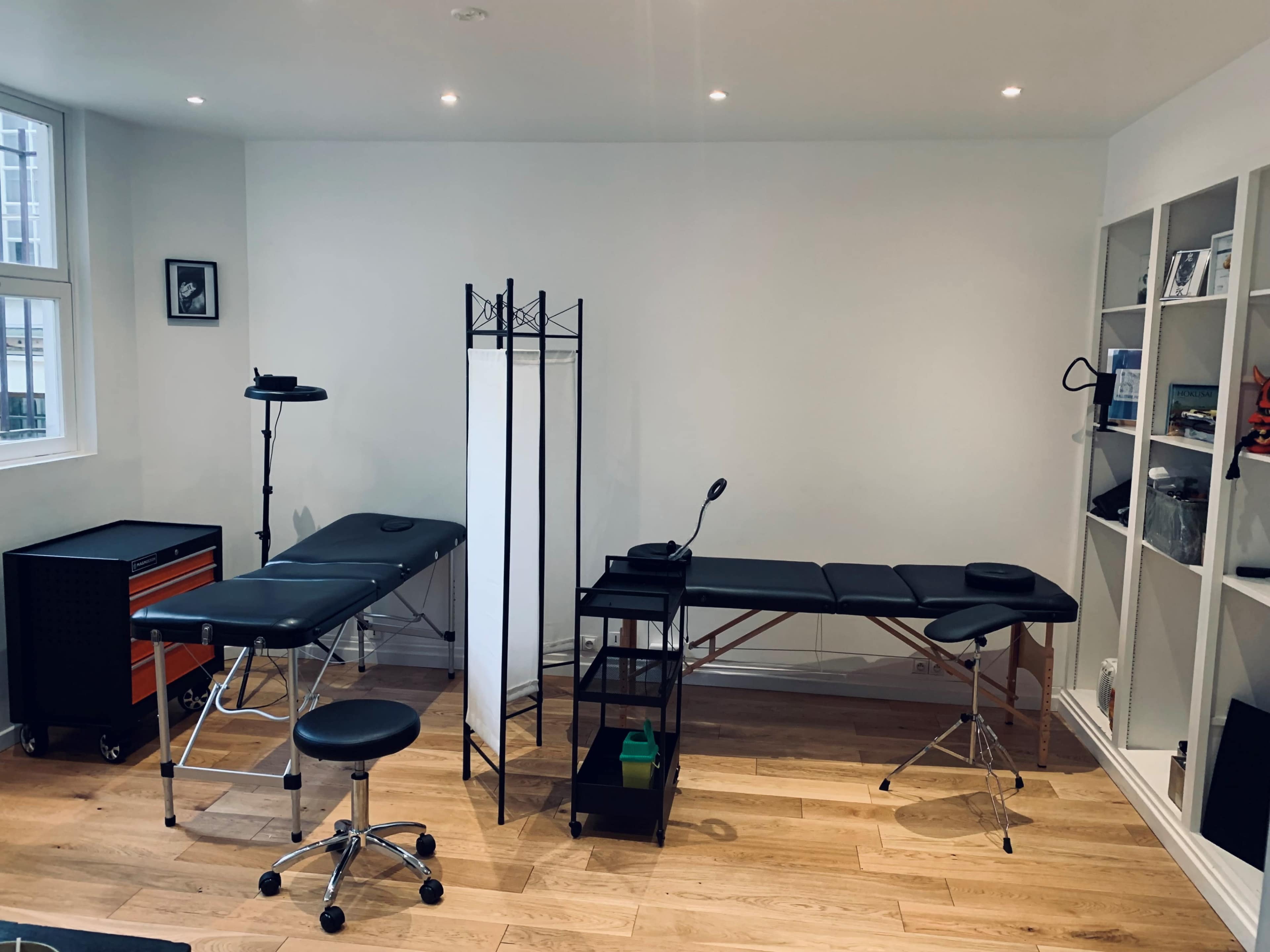 A minimalist treatment room featuring two examination tables, a privacy screen, a task light, and a shelving unit against a white wall.