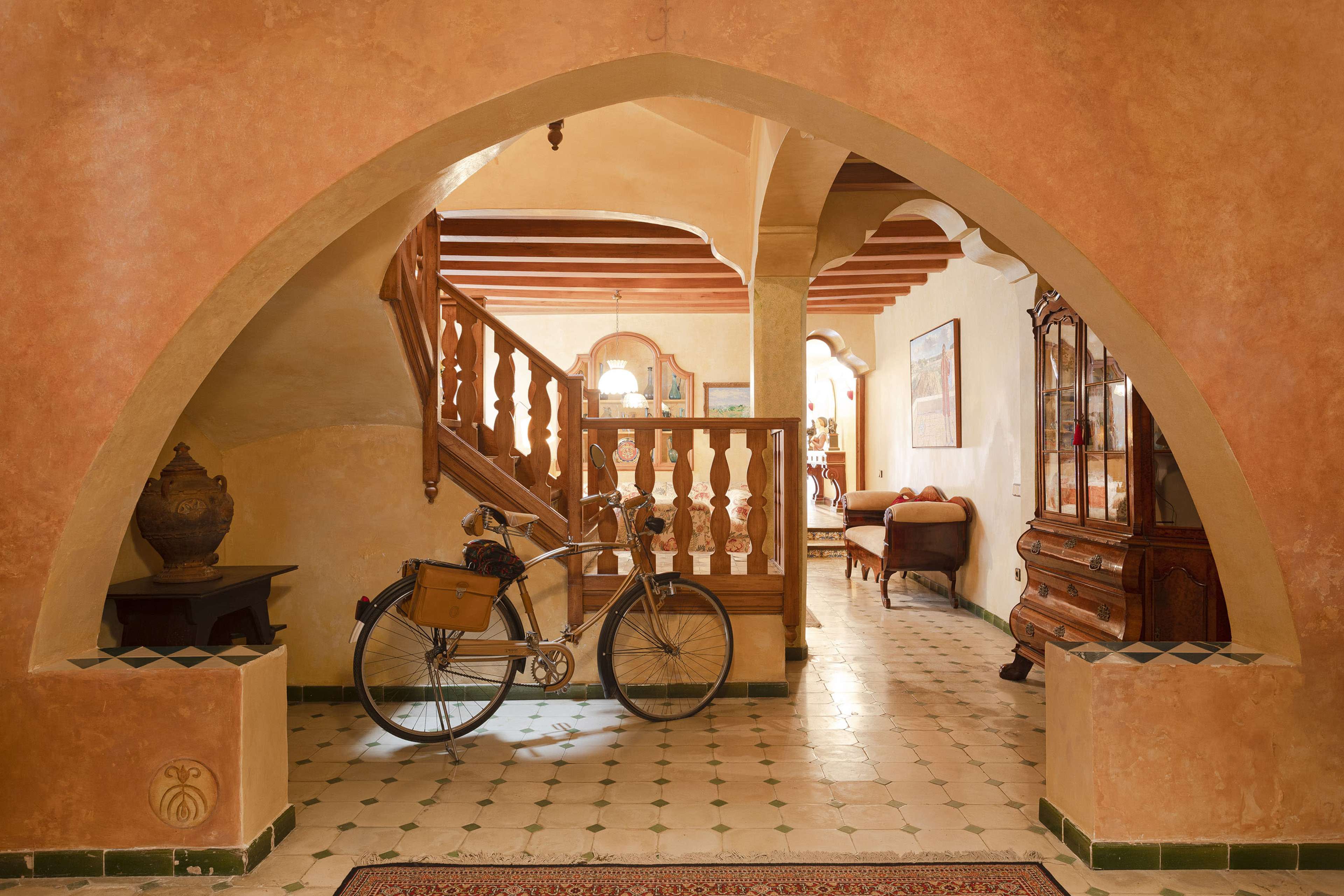 A well-decorated hallway with a bicycle parked by an arch, leading to a staircase and a seating area in a warm-toned home interior.