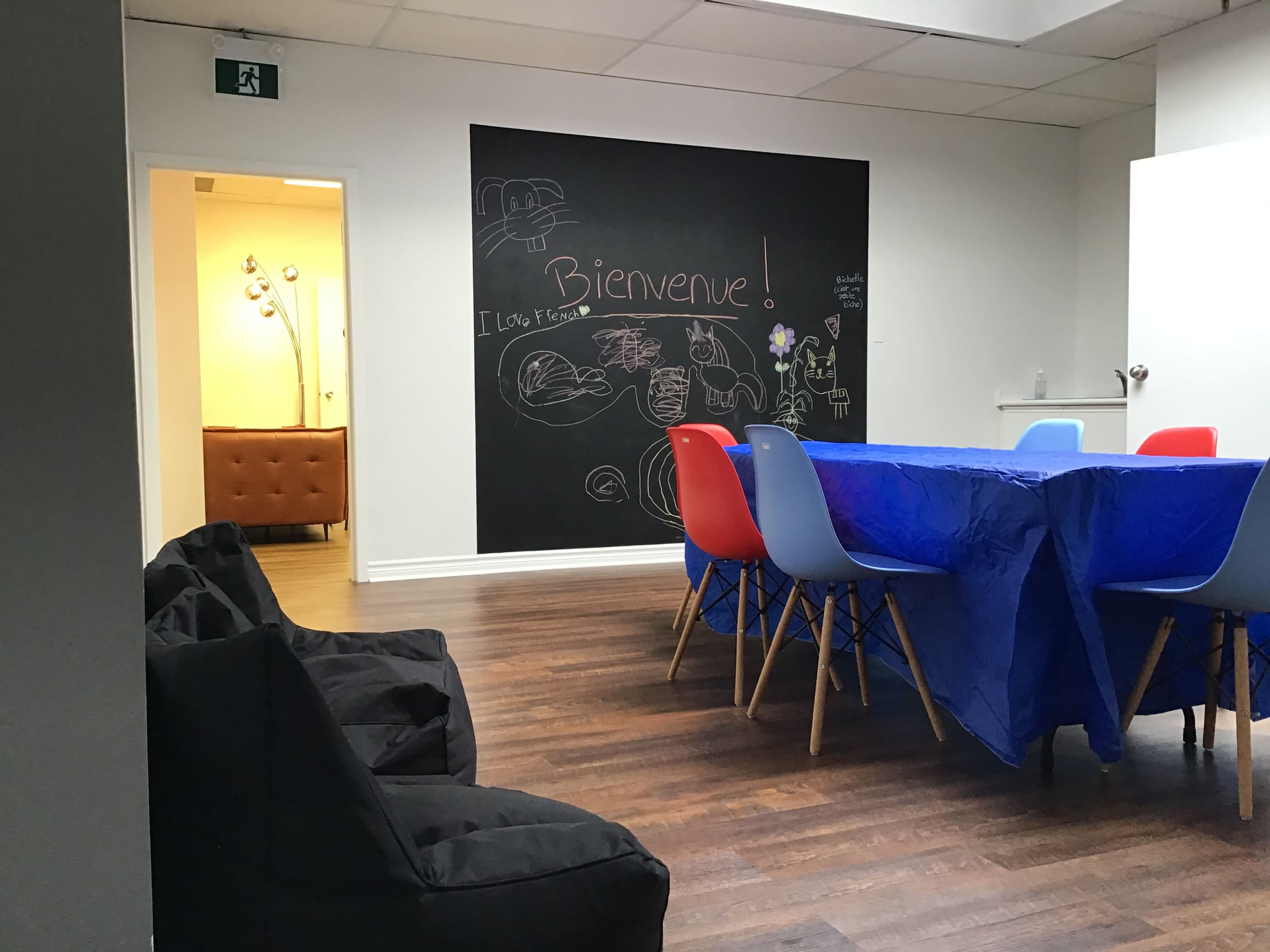 A room with a large blue table surrounded by colorful chairs, a chalkboard wall with drawings and text, and a cozy seating area in the background.