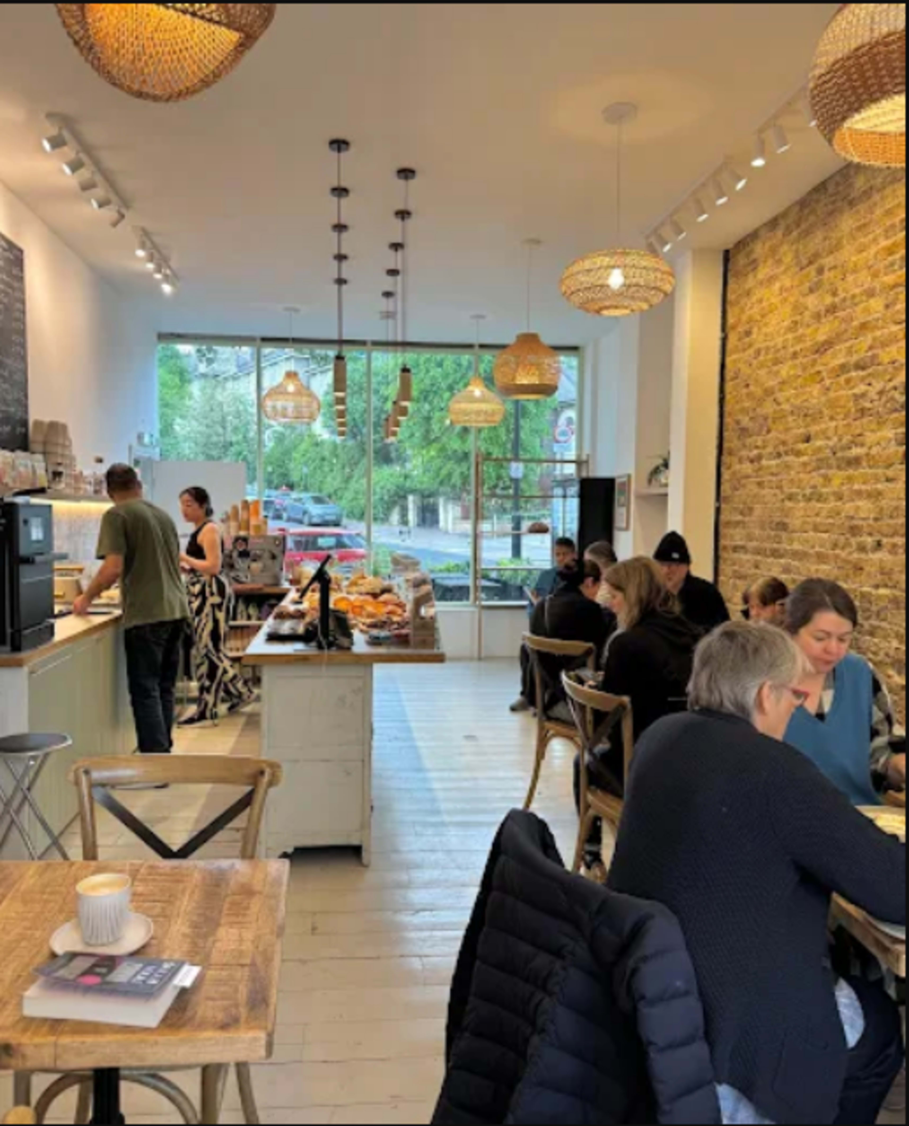 A café interior features customers seated at wooden tables, with a barista preparing drinks behind the counter, under hanging light fixtures.