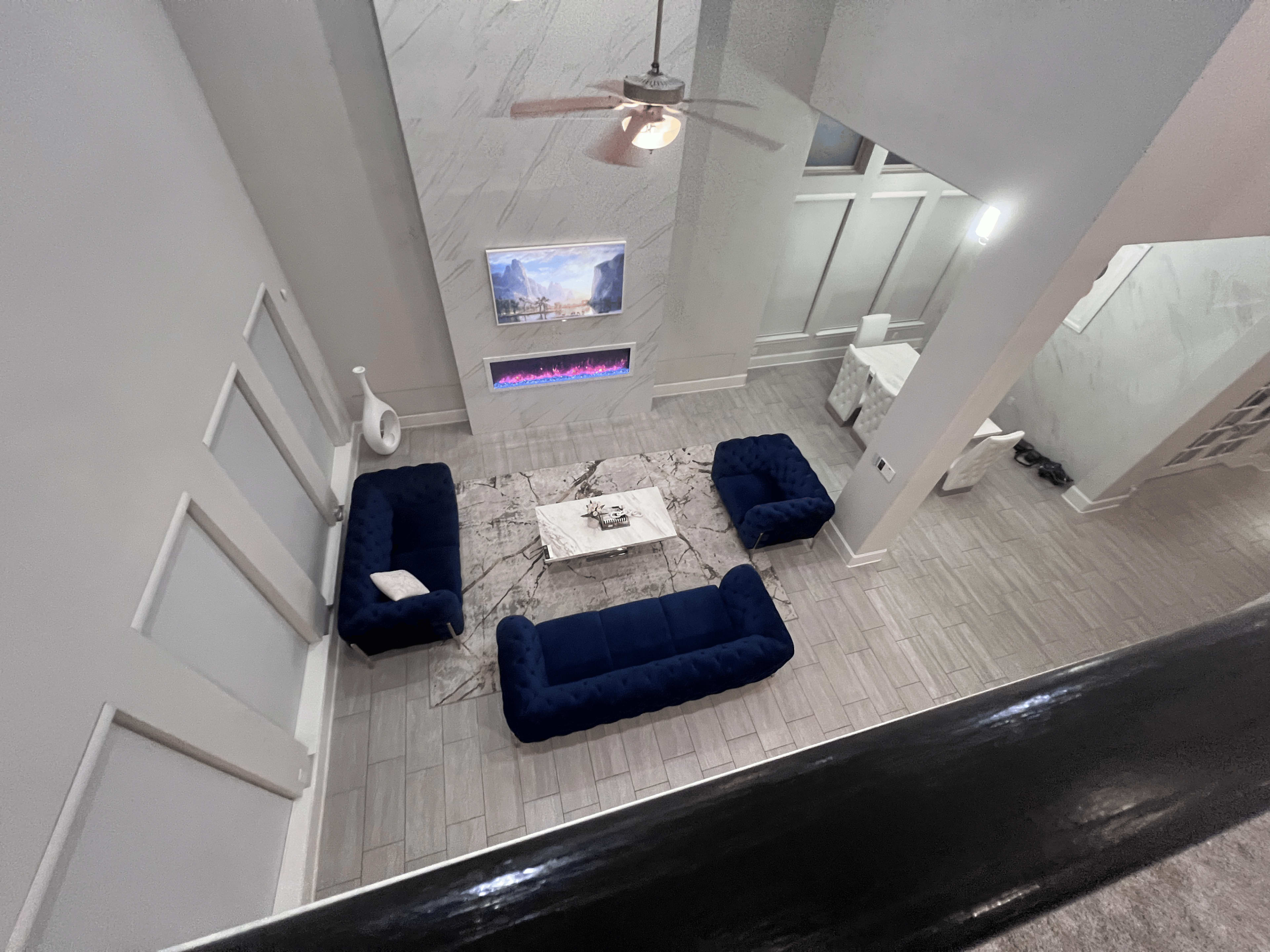 The image shows a modern living room featuring two dark blue sofas arranged facing a coffee table, with a mounted TV and an electric fireplace on the wall.