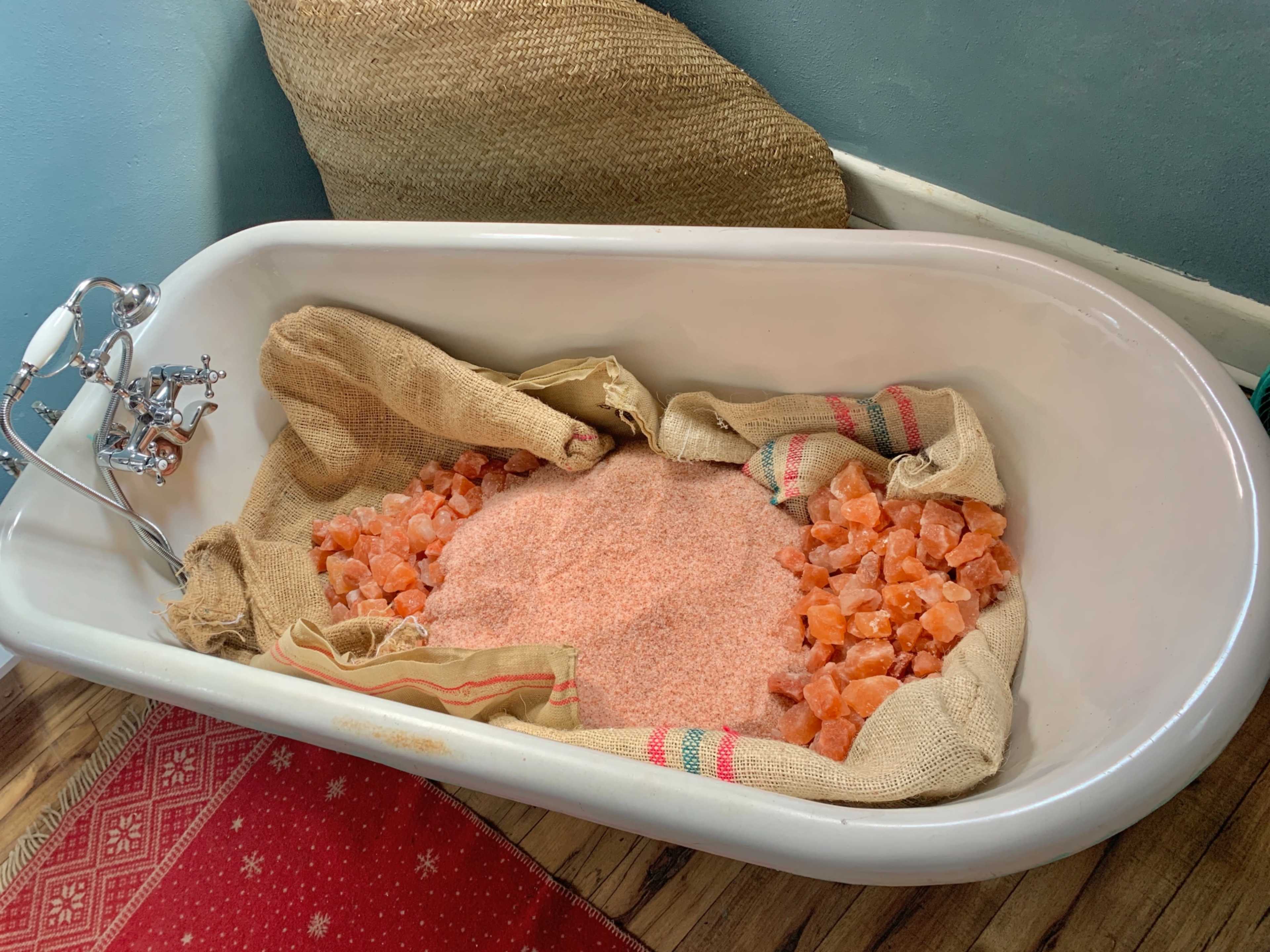 A vintage bathtub is filled with pink Himalayan salt, surrounded by burlap sacks and placed on a wooden floor with a patterned rug.
