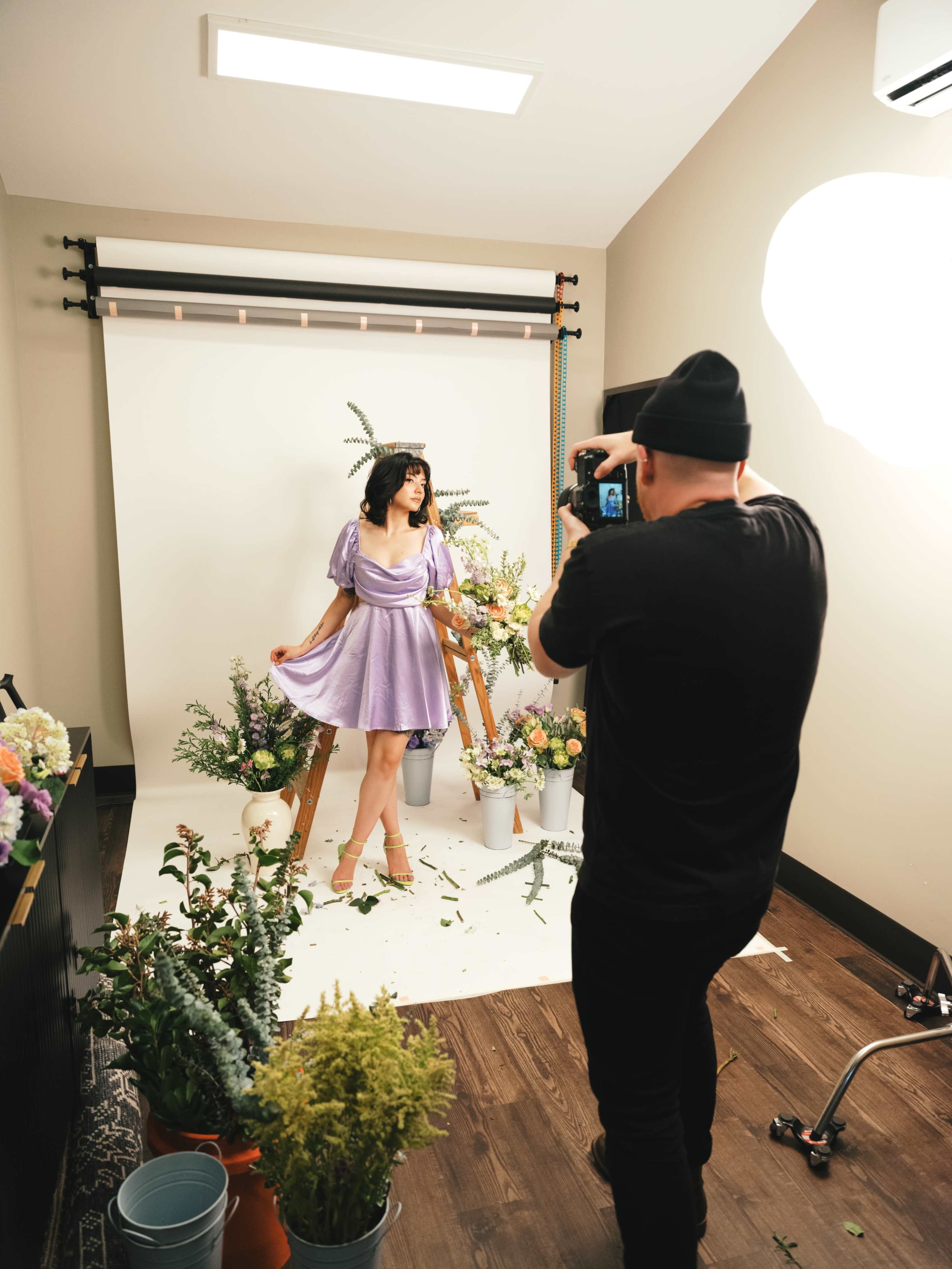 A person in a purple dress poses in front of a backdrop while another person takes photos with a camera.
