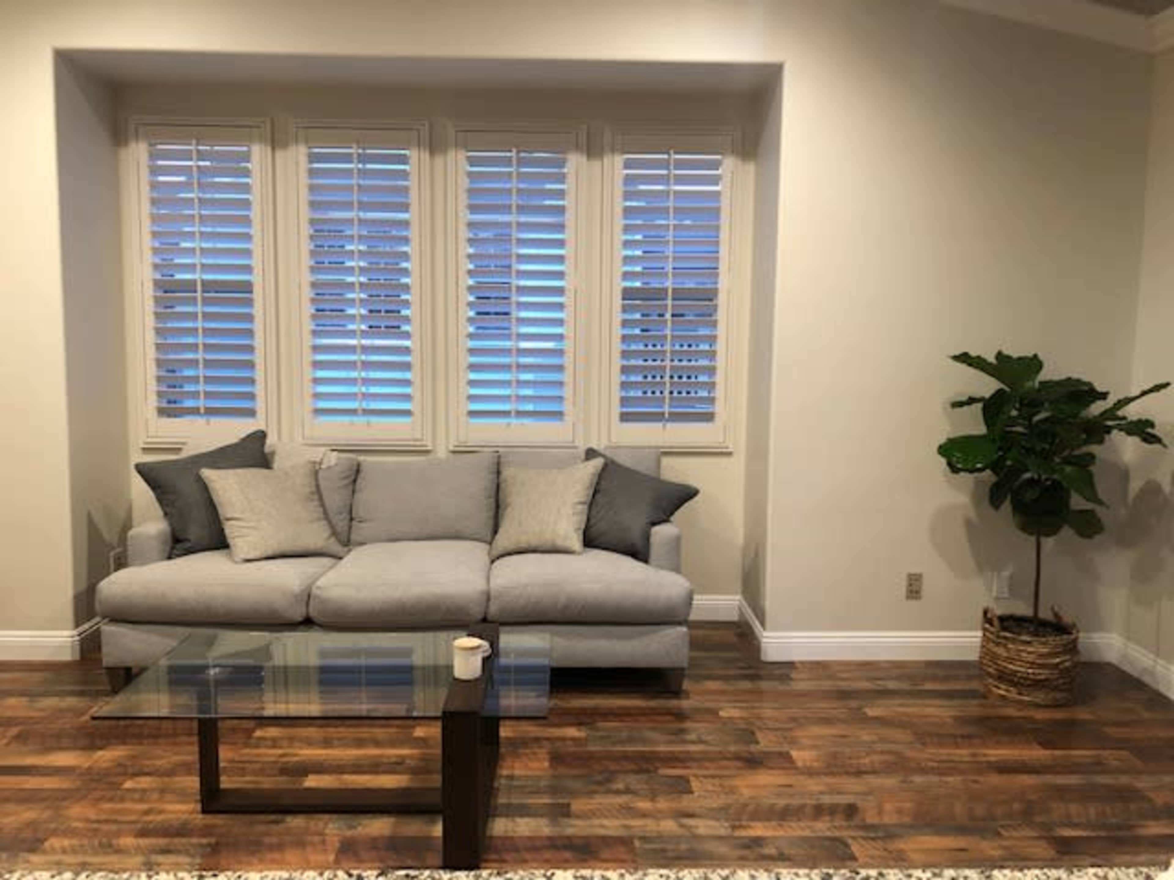 The image shows a light gray sofa with several cushions, positioned in front of a window with white shutters, along with a glass coffee table and a potted plant in a woven basket.