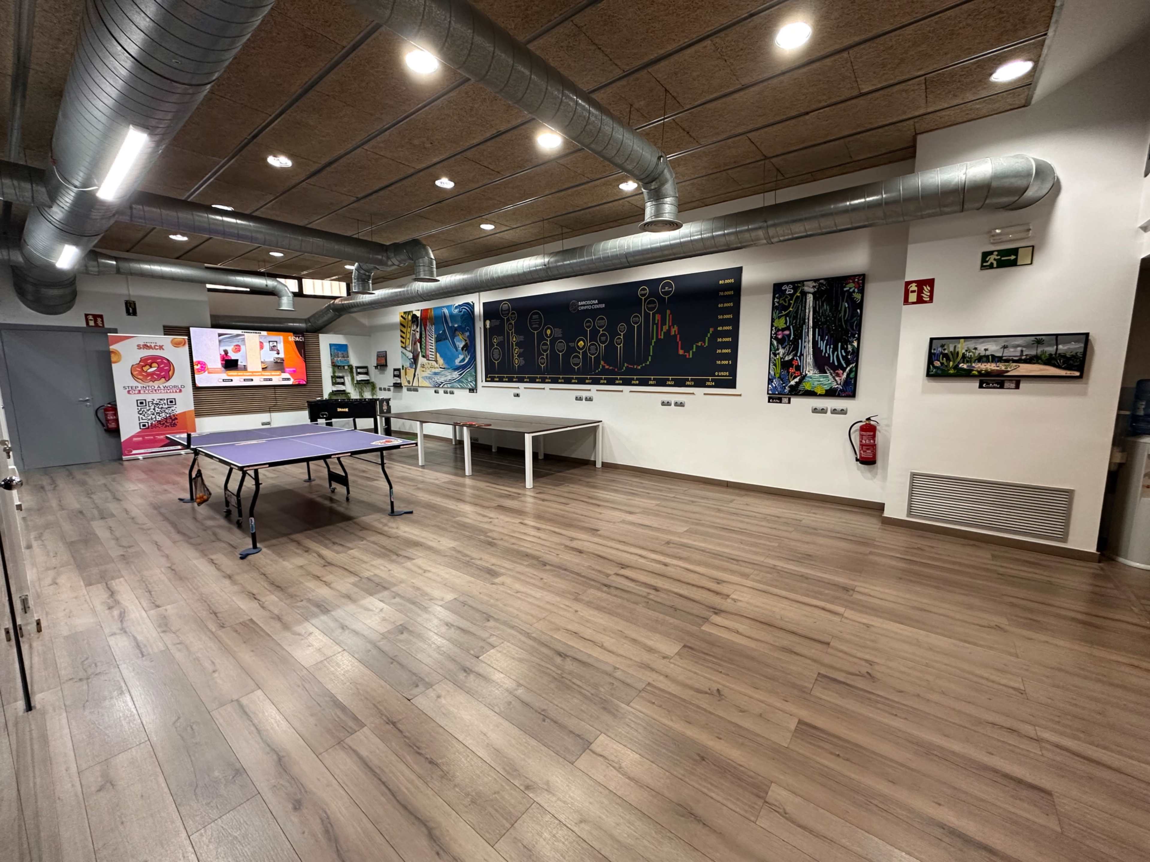 The image shows a spacious room with wooden flooring, featuring a ping pong table, a white table, and colorful artwork displayed on the walls.