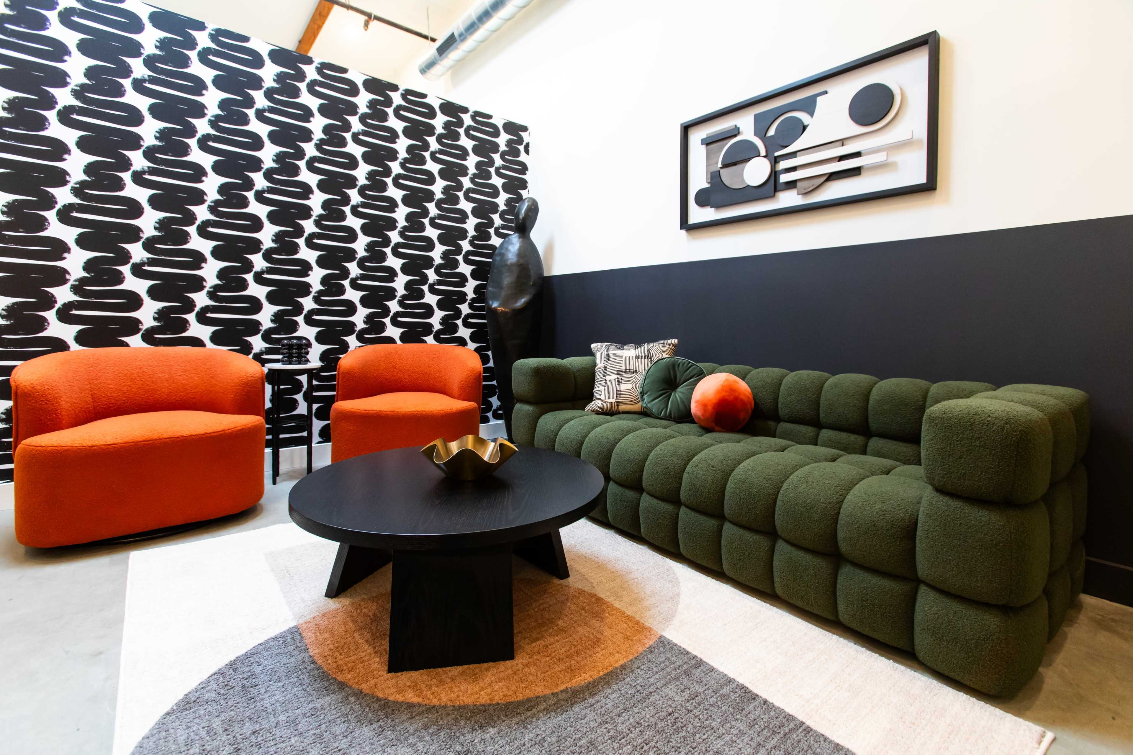 A living room with a green tufted sofa, two orange armchairs, a black round coffee table, and an abstract wall art piece against a patterned black and white backdrop.