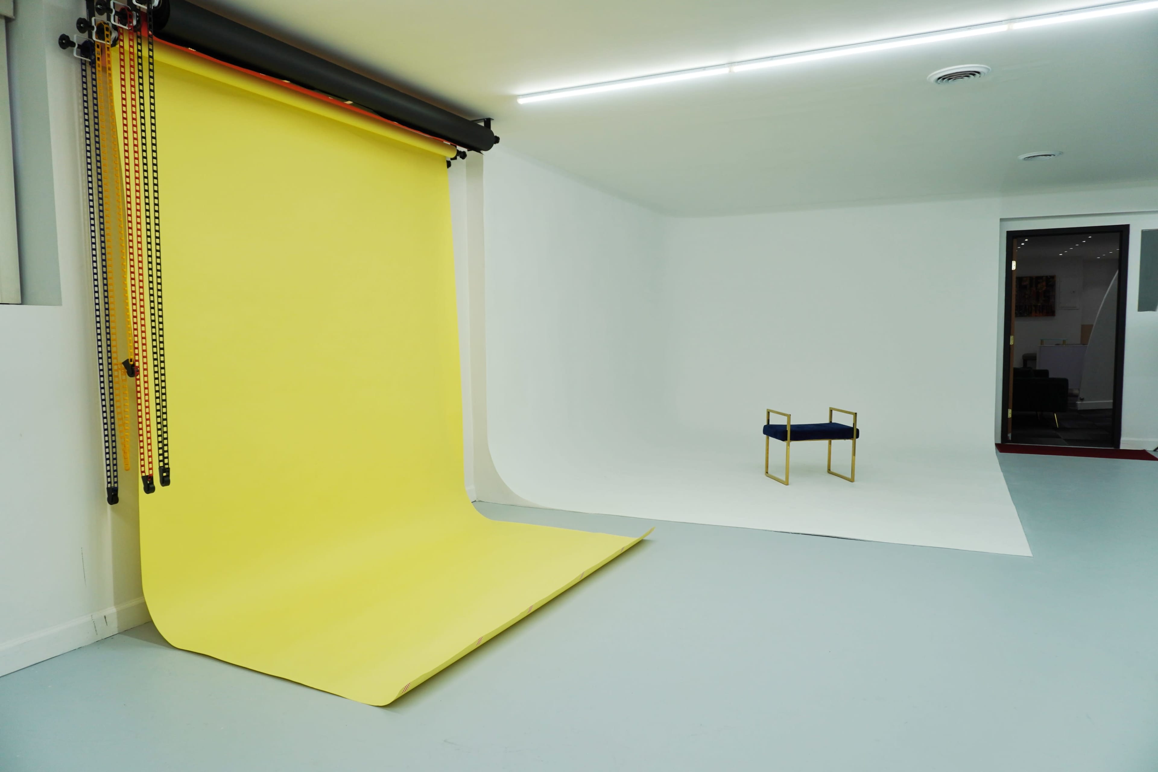 The image shows a minimalistic photography studio with a yellow backdrop on one side and a white backdrop on the other, featuring a single blue chair placed in front of the white background.