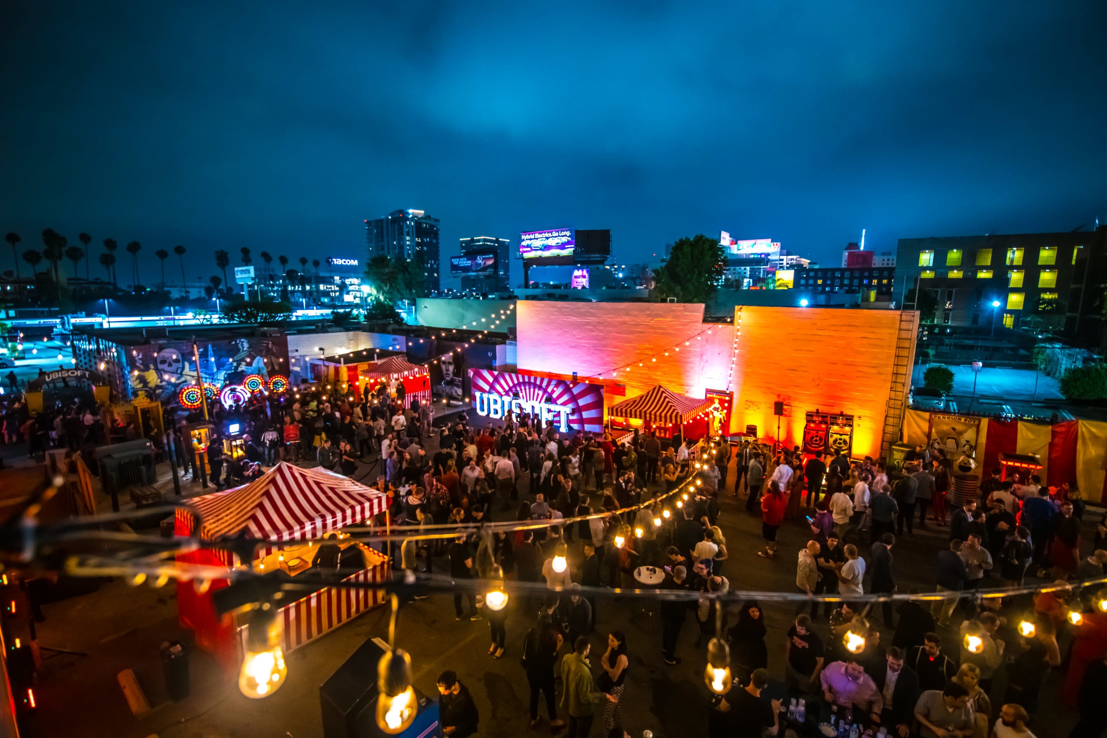 A rooftop event space is filled with people enjoying festivities under string lights and colorful decorations, with a skyline view in the background.