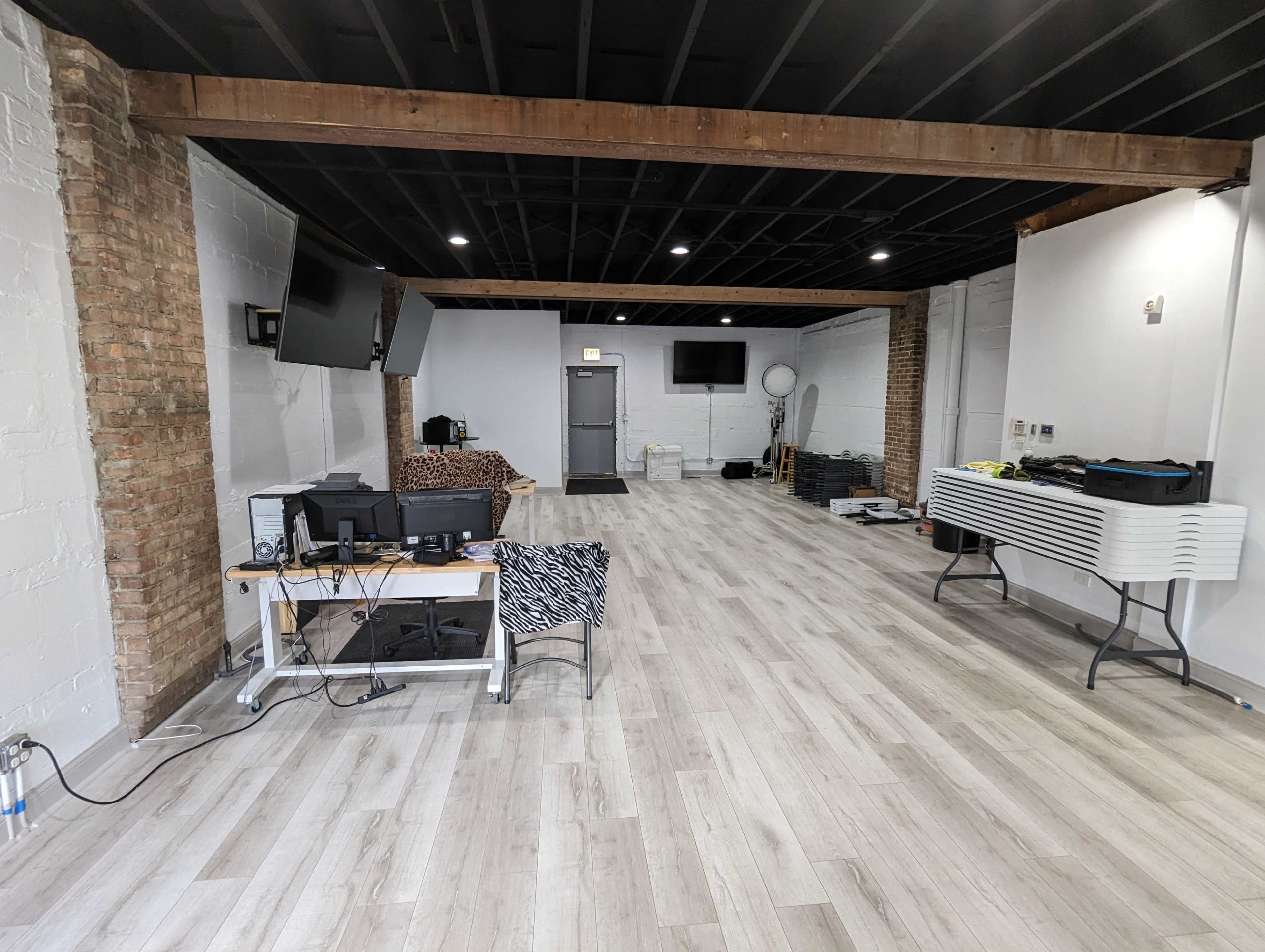 An empty interior space with wooden beams, two mounted televisions, a desk with computer equipment, and a stack of equipment along the wall.