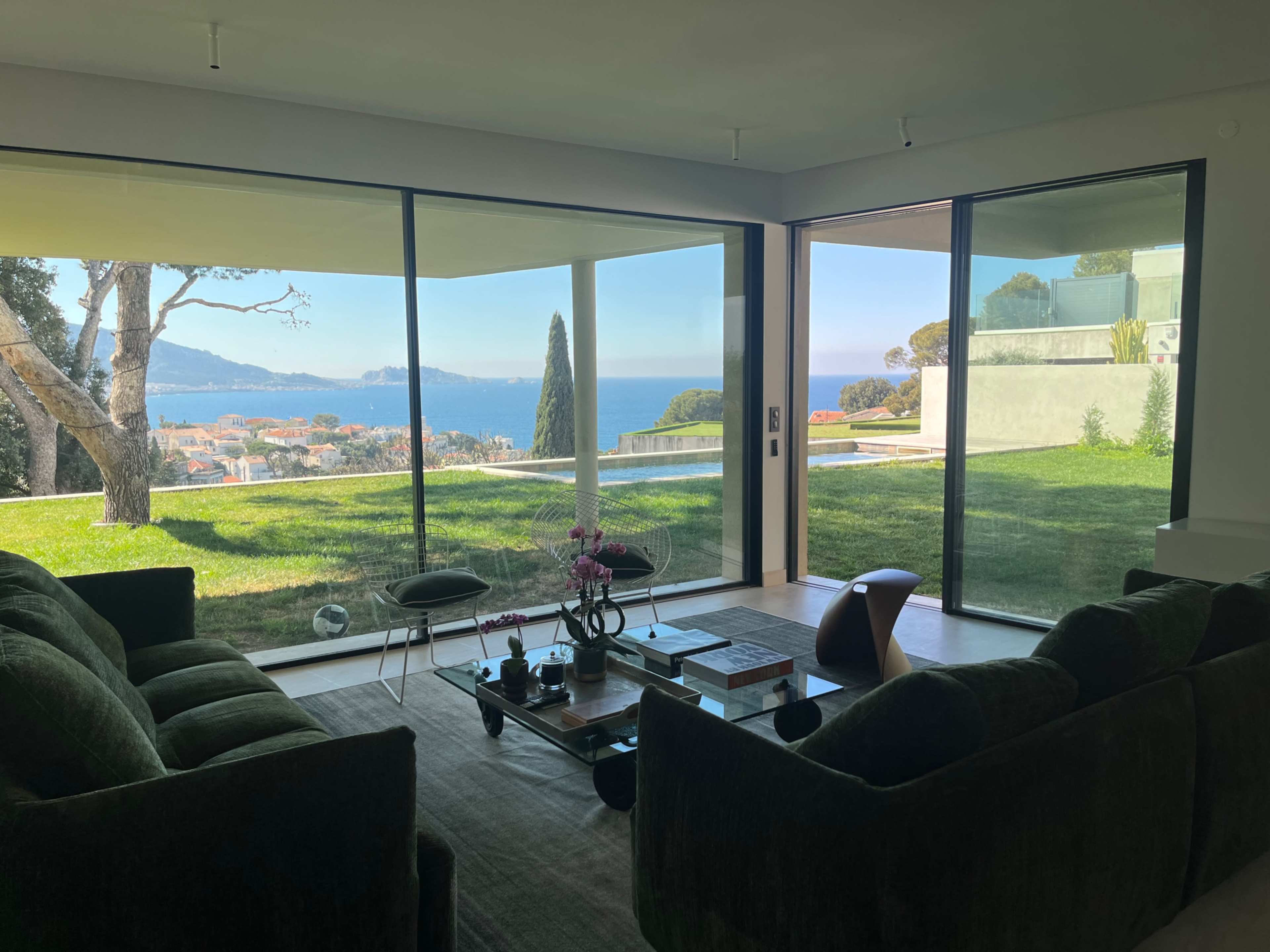 The scene shows a living room with large windows that overlook a scenic view of the sea and surrounding landscape.