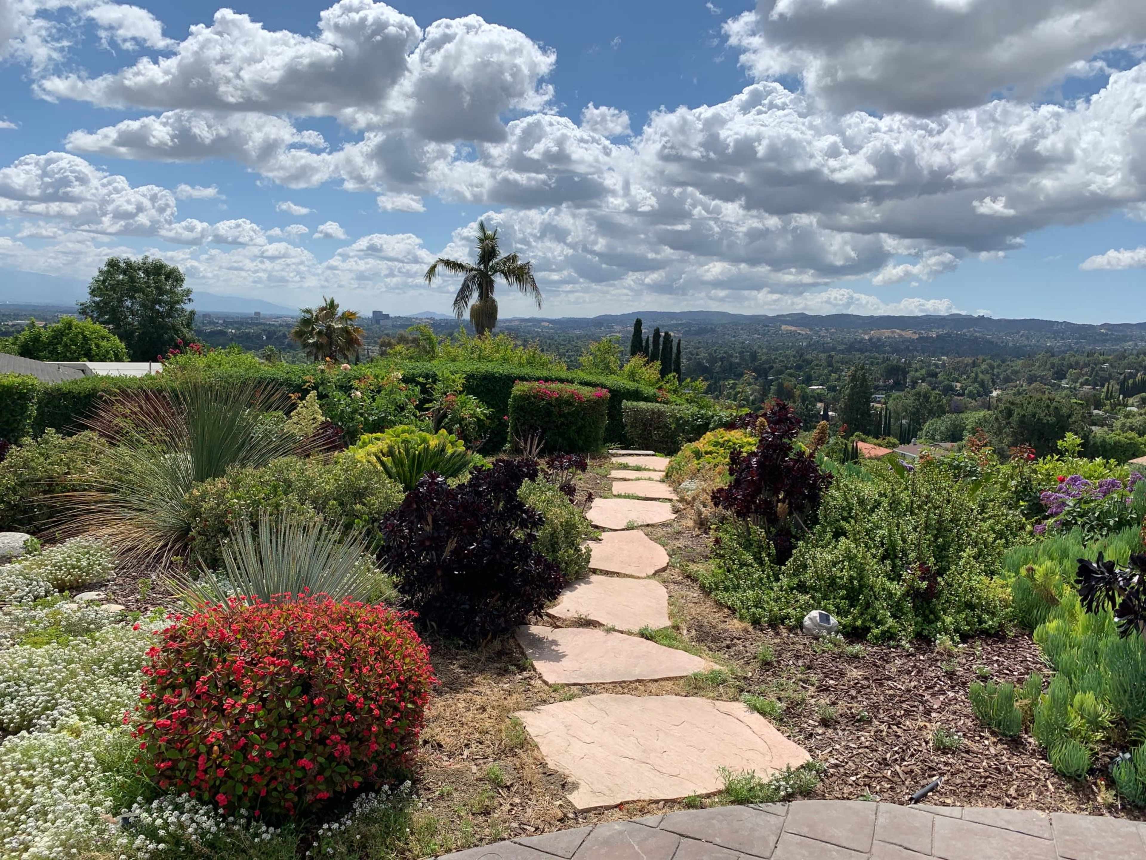 The image depicts a stone pathway winding through a lush garden with colorful flowers and greenery, set against a backdrop of clouds and distant hills.