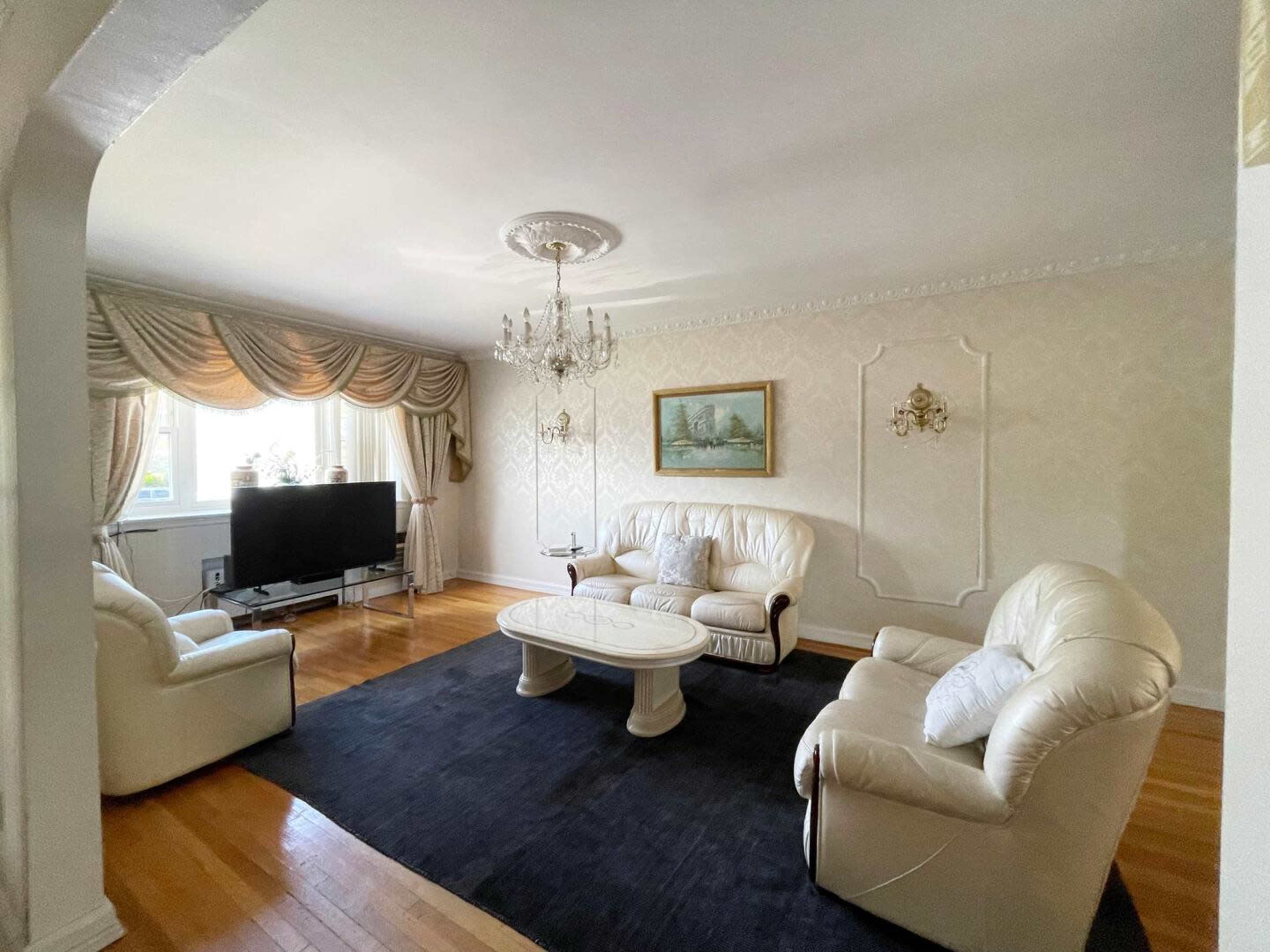 The living room features white leather furniture arranged around a black area rug, with a television, chandelier, and decorative wall elements.