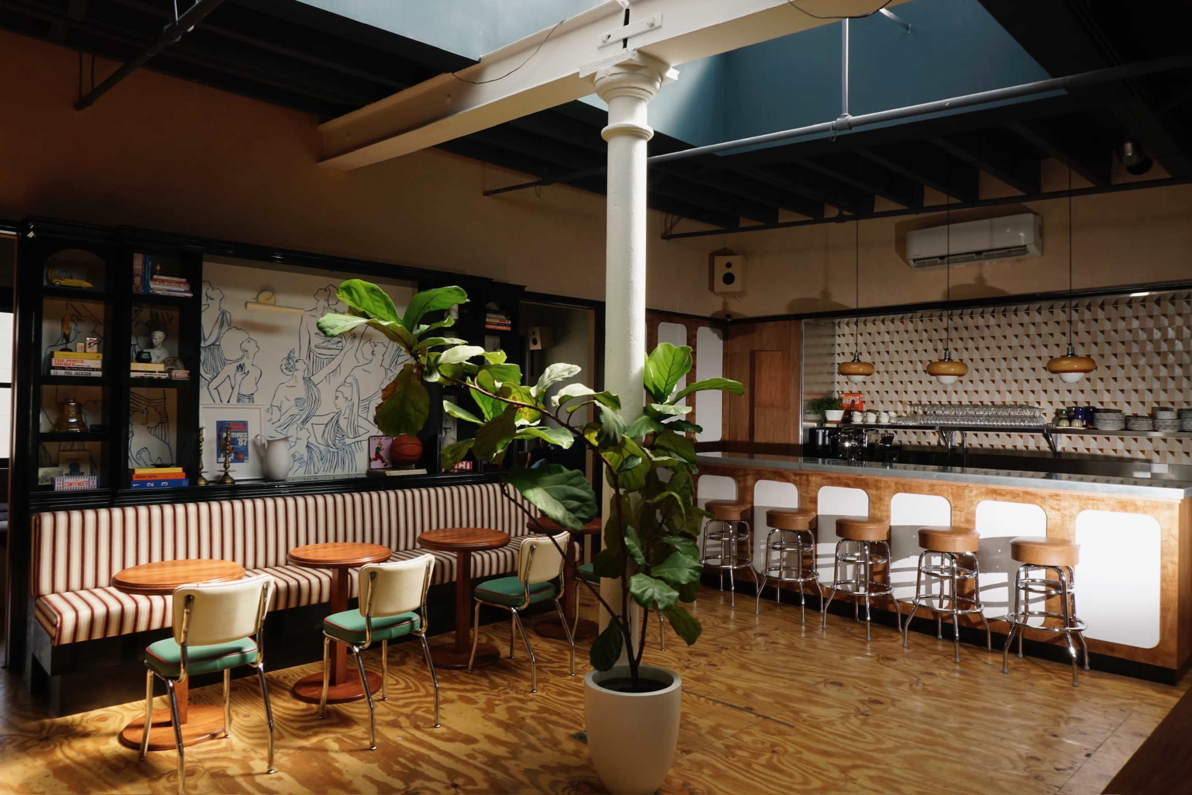 The image shows a spacious café interior with striped seating, circular tables, a bar area, and large windows letting in natural light.