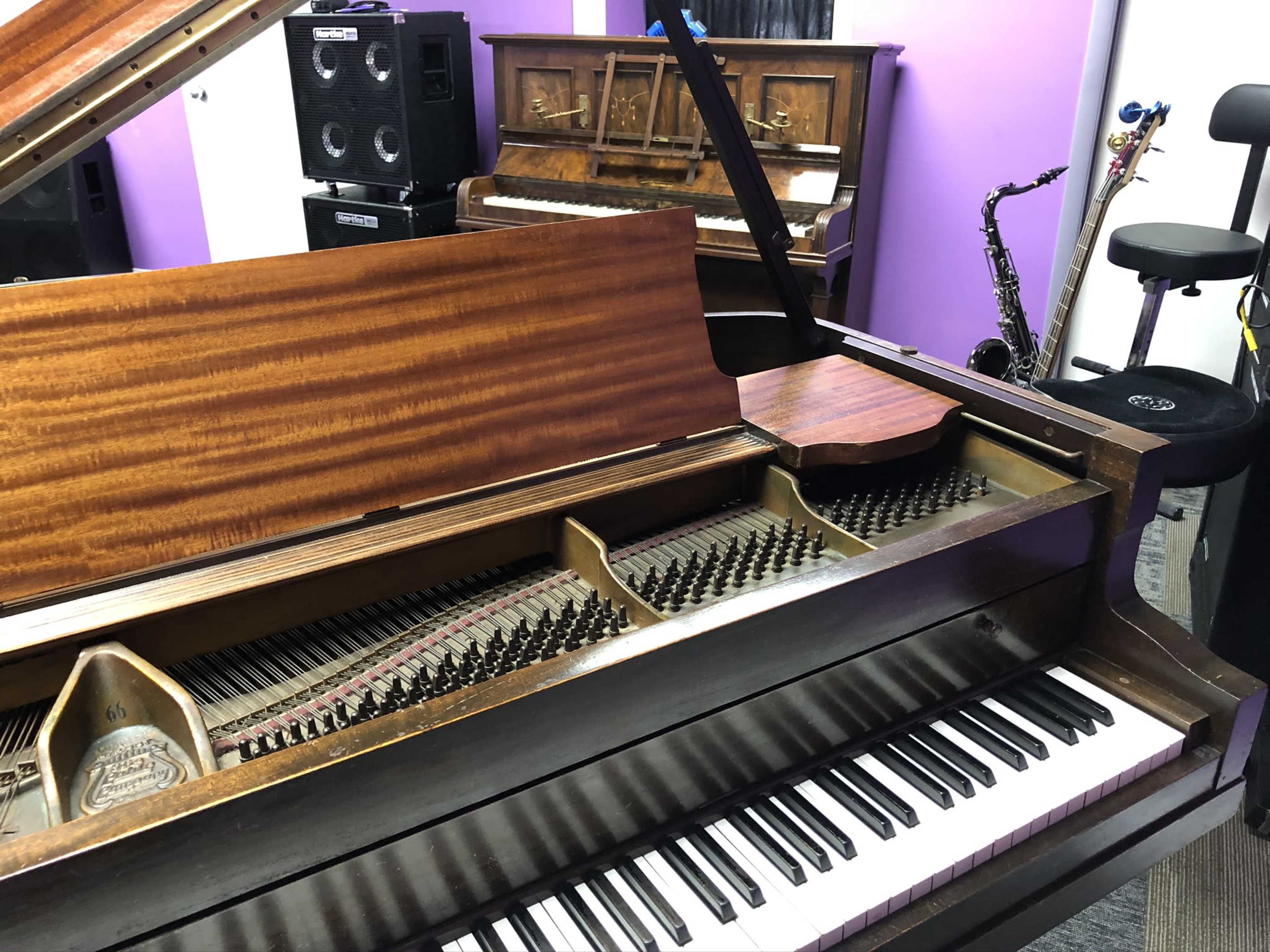 The interior of an open grand piano, revealing its strings and hammers, with two additional pianos and a saxophone in the background.