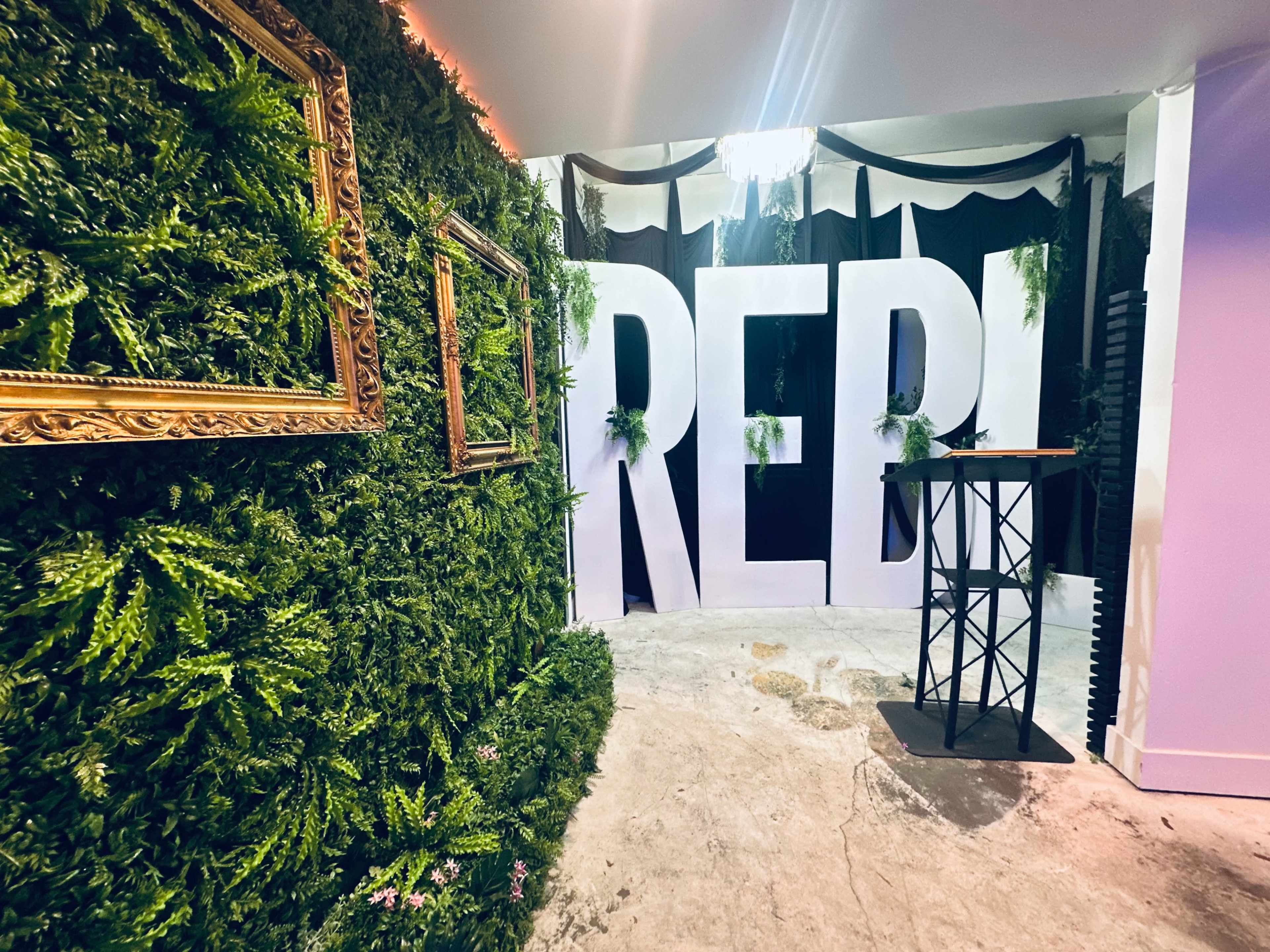 The image shows a decoratively designed space featuring a green wall with a framed mirror and a large white sign that reads "REBI," along with a black podium for events.