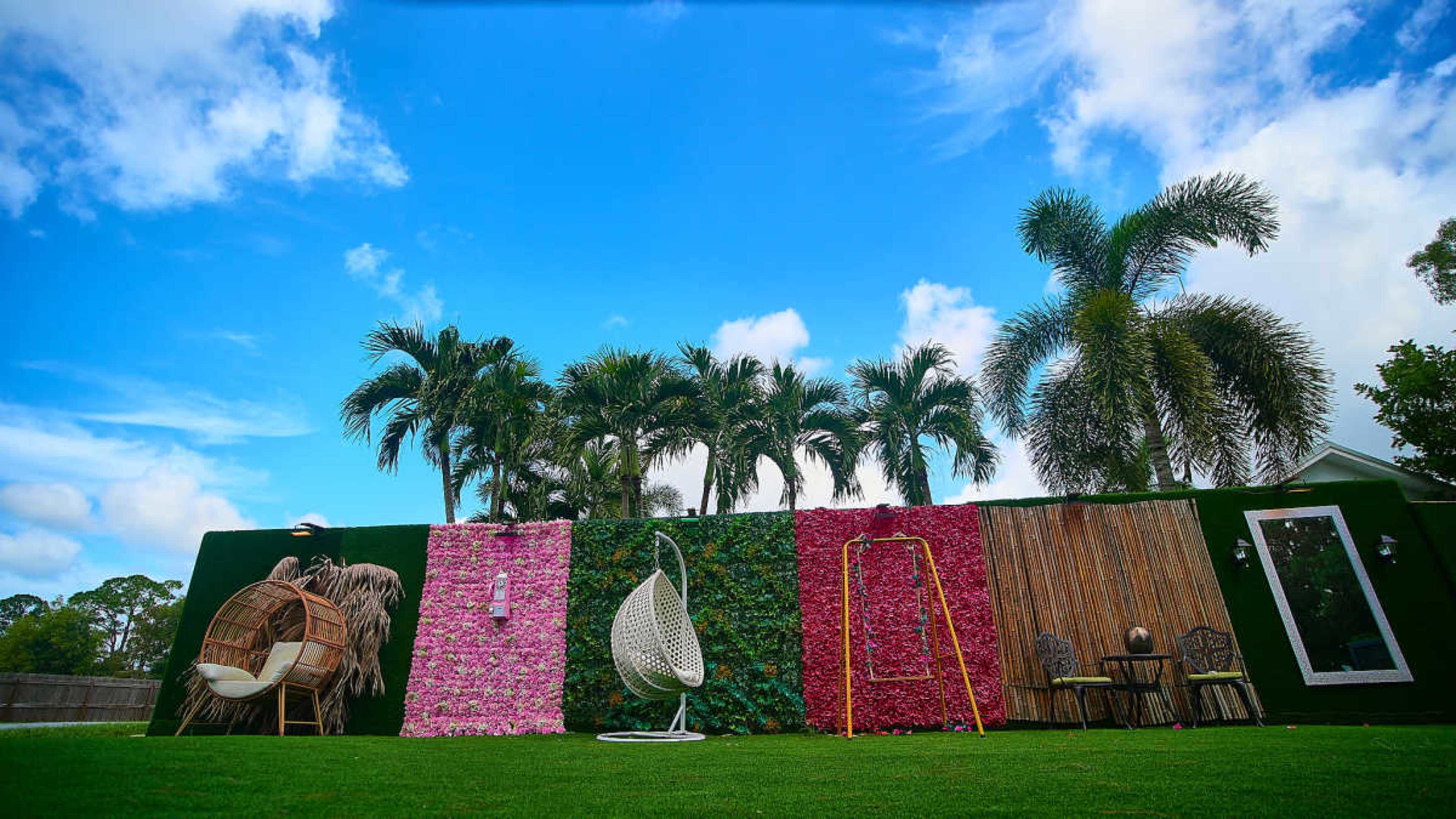 The image shows a landscaped outdoor area featuring two hanging chairs, a colorful wall adorned with flowers, and palm trees under a blue sky.