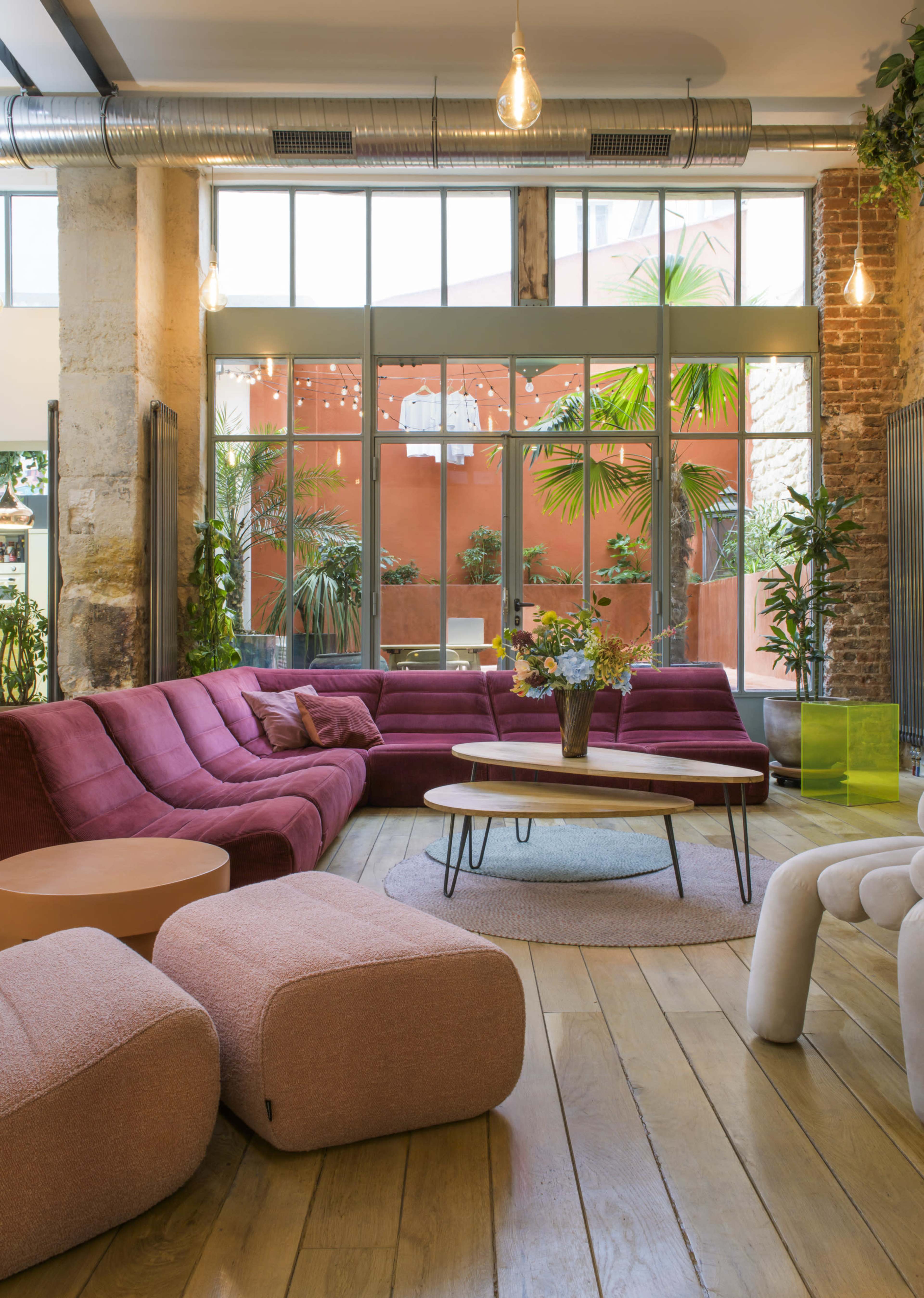 A modern living area features a large pink sectional sofa, a round coffee table, and potted plants against a backdrop of large windows and red walls.