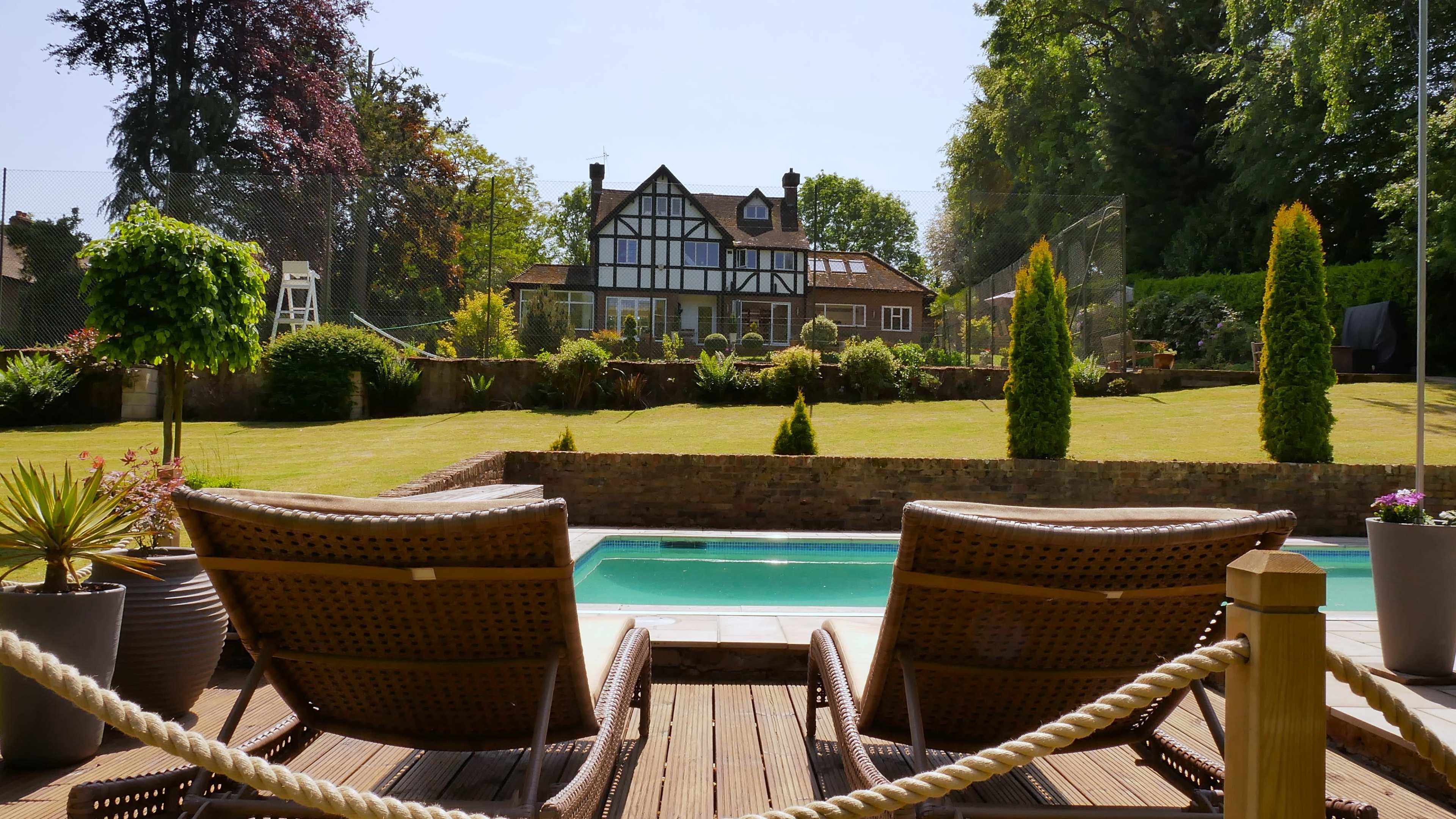 A landscaped backyard features a swimming pool in front of a large, two-story house with a timber-framed design.