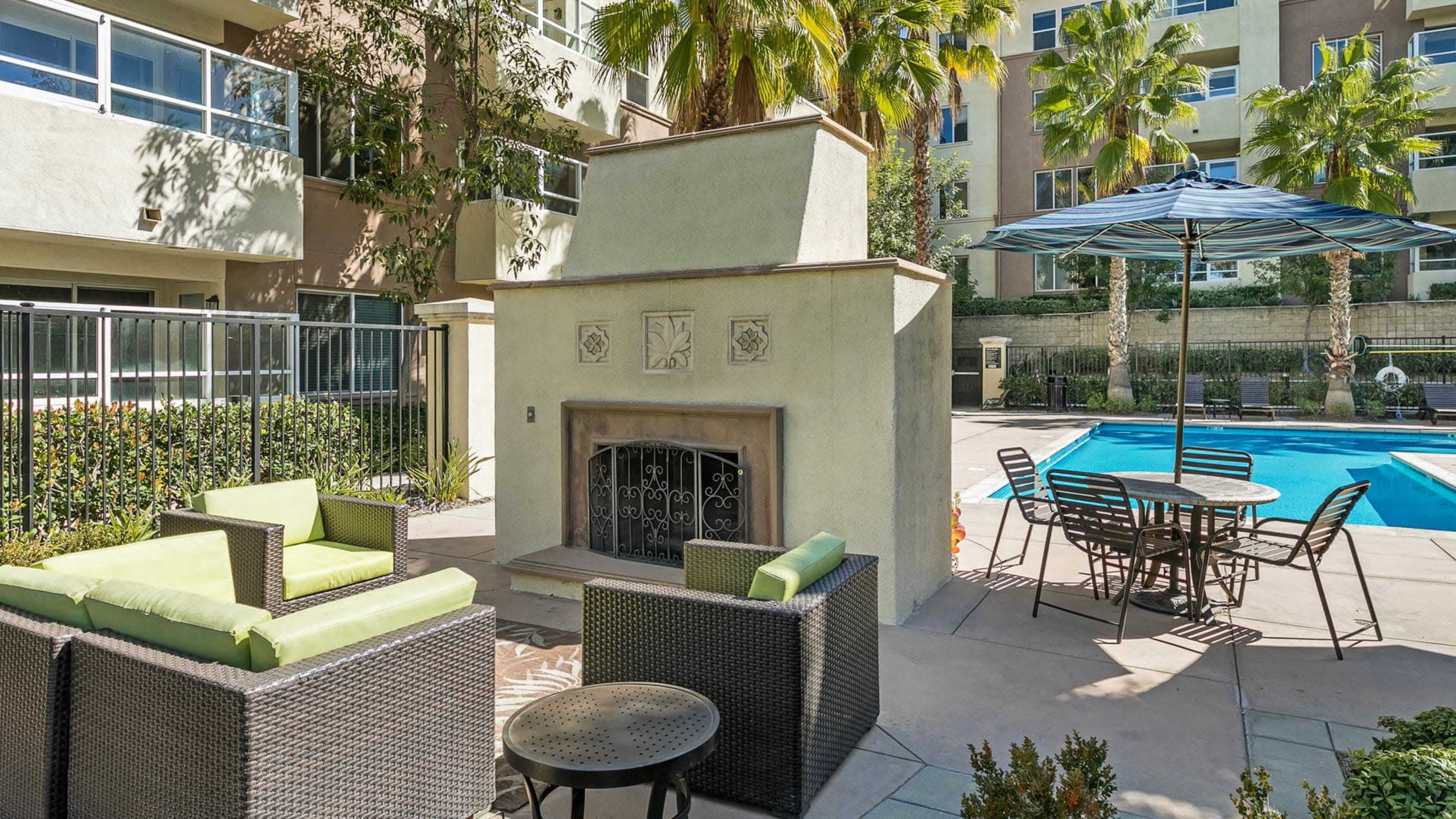 A seating area with green chairs is located near a swimming pool, featuring a fireplace and umbrellas surrounded by palm trees.