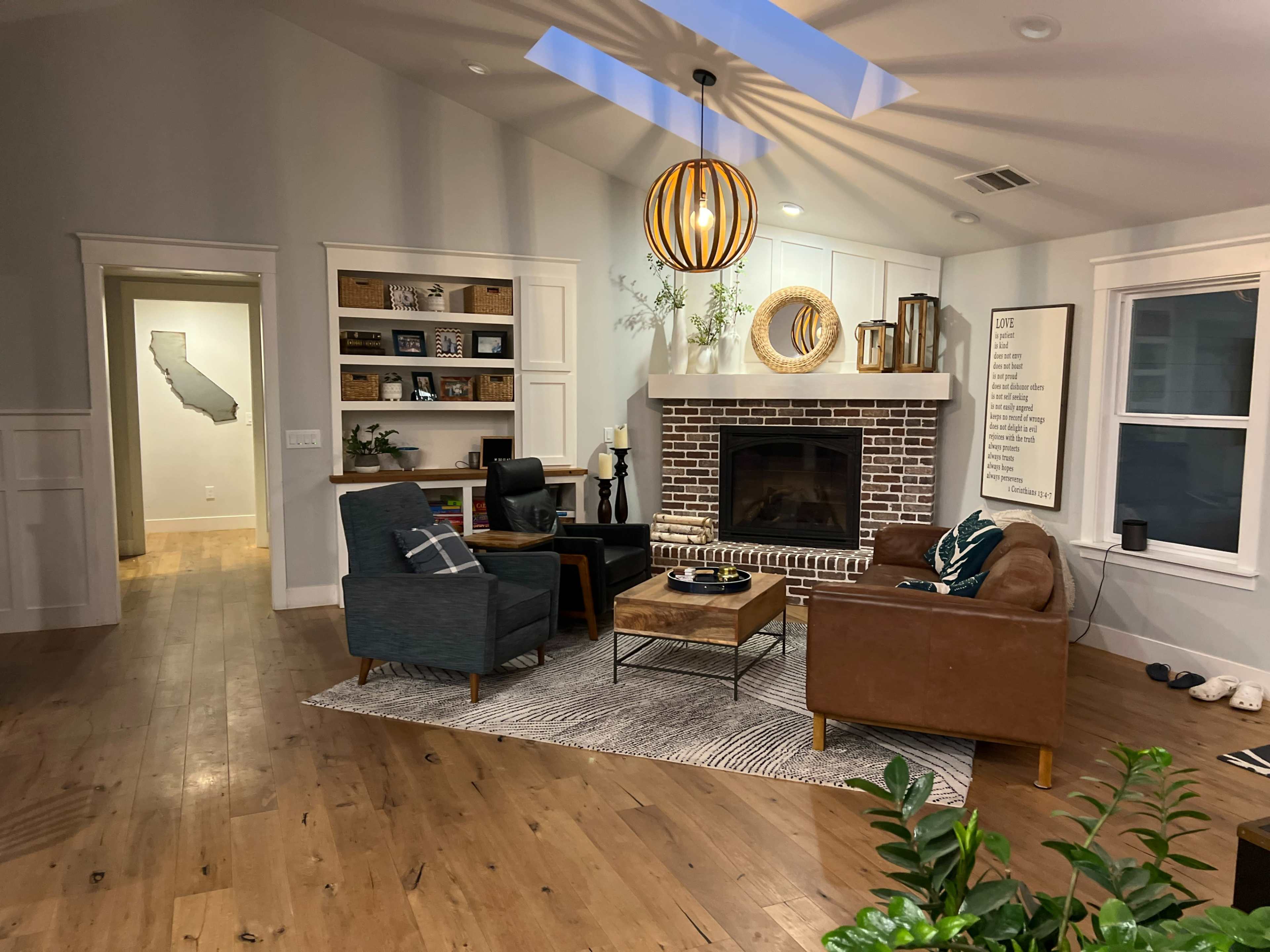 A cozy living room features a brick fireplace, two armchairs, a coffee table, and a large wall mirror, with a doorway leading to another room.
