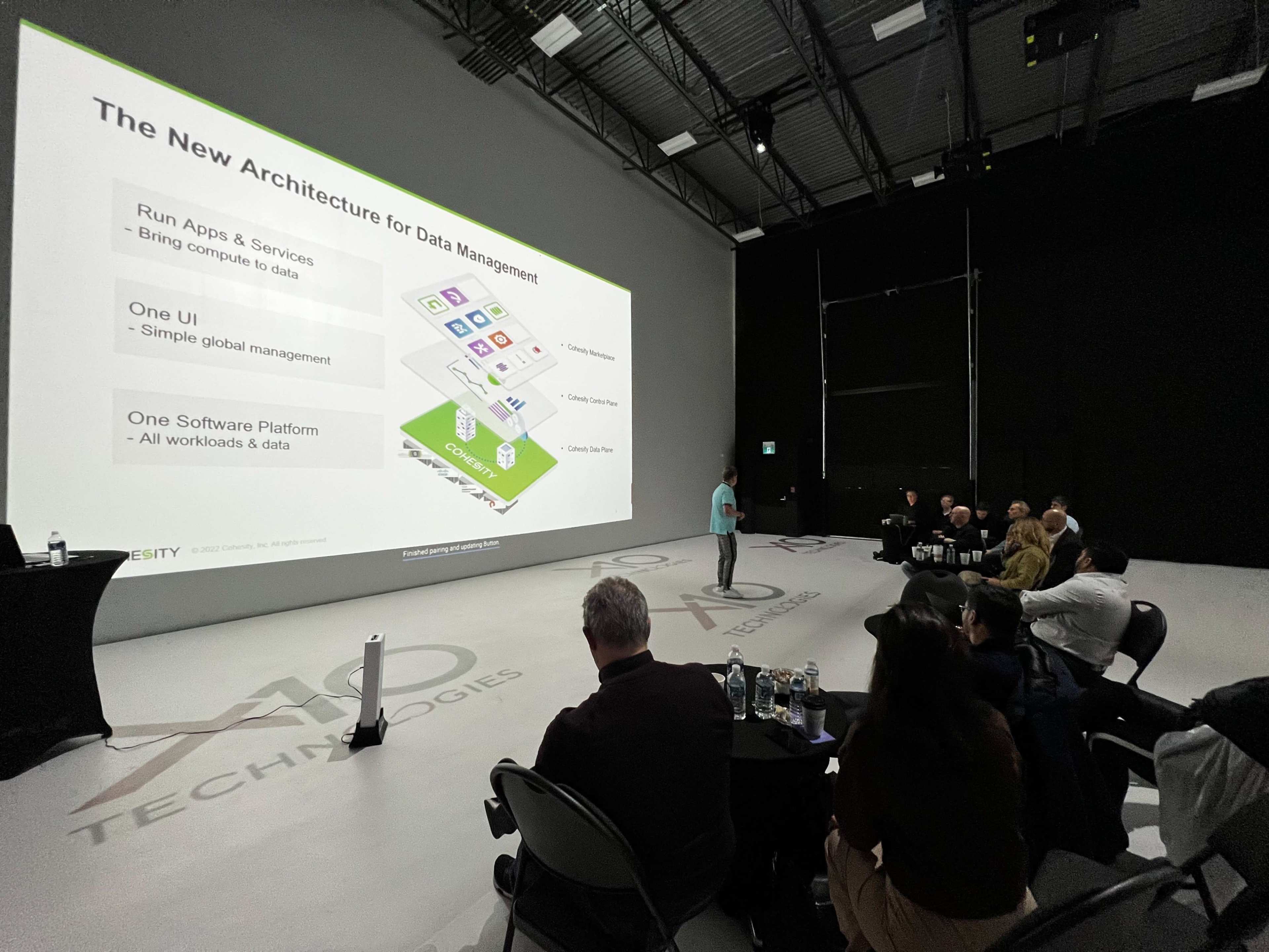 A presenter stands in front of a large screen discussing a new architecture for data management to an audience seated in a studio.