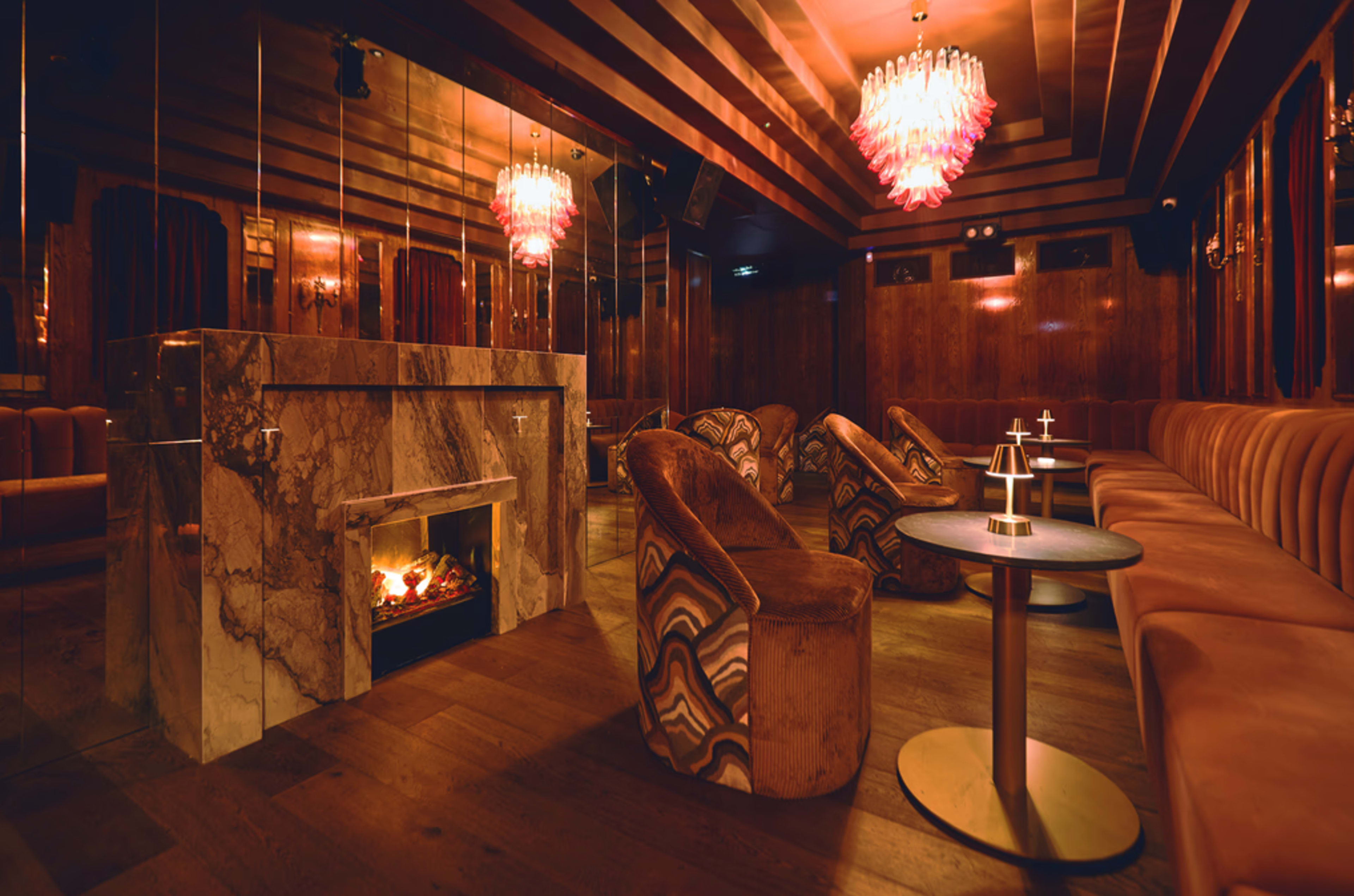 The image shows a stylish lounge with plush seating, a marble fireplace, and decorative chandeliers.