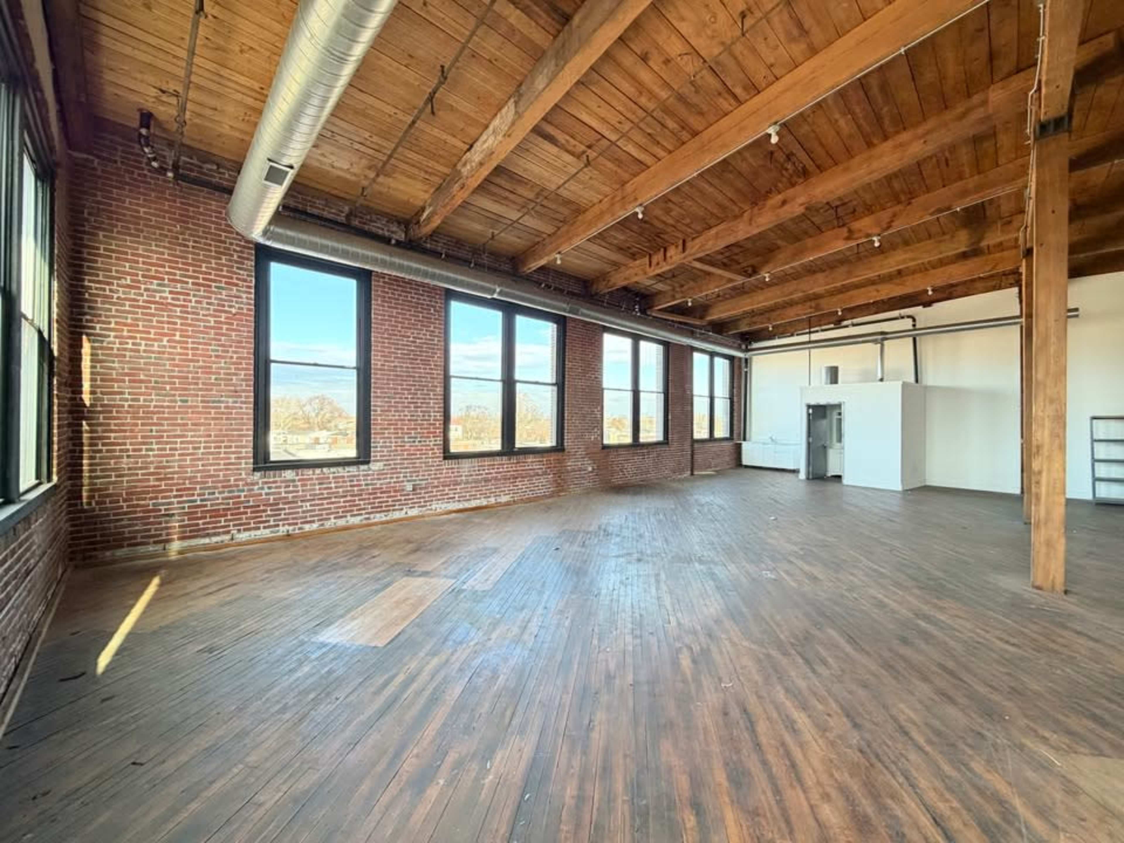 The image shows a spacious, sunlit room with large windows, exposed brick walls, and wooden beams on the ceiling.