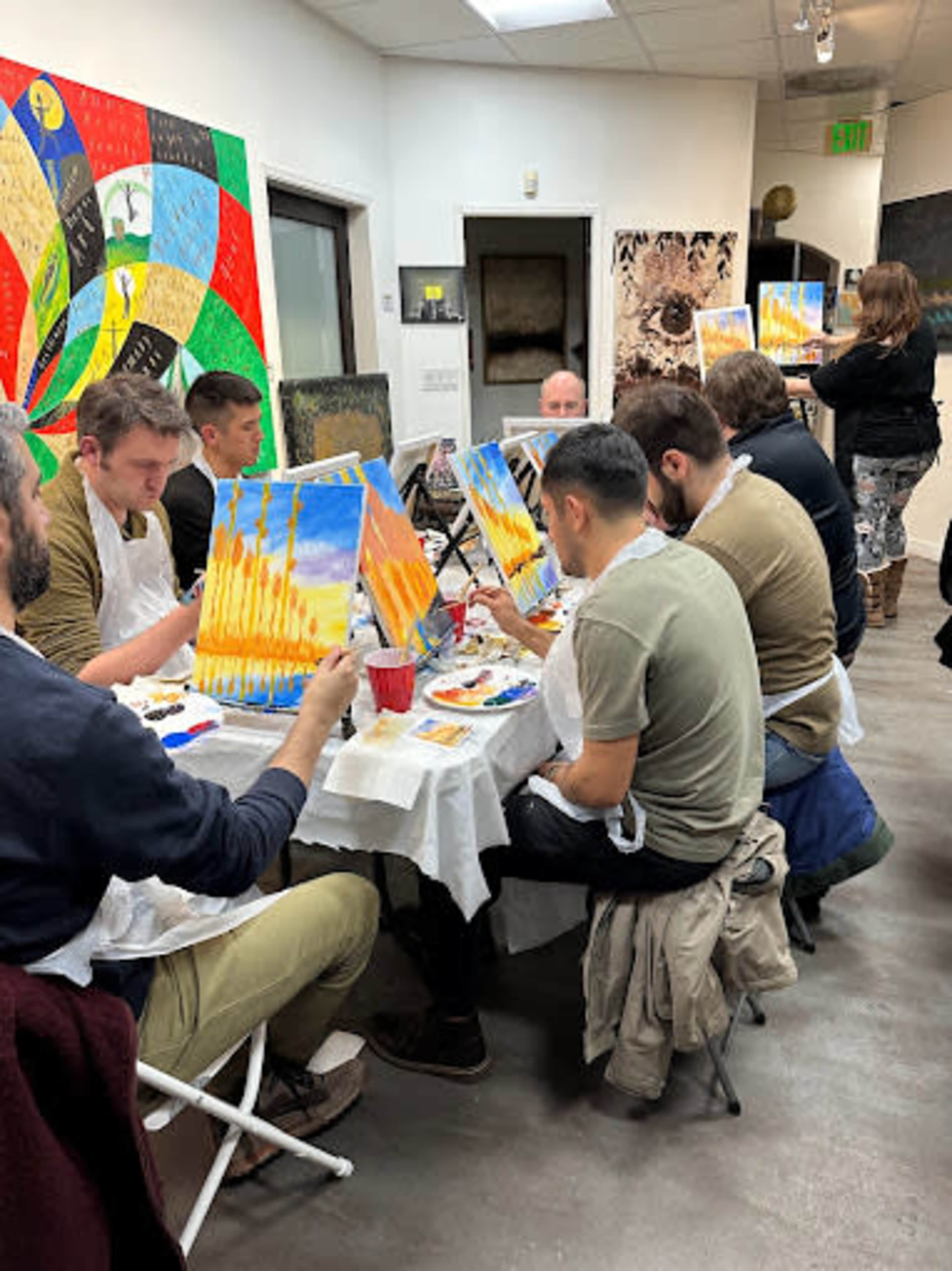 A group of people sit at tables in an art studio, each painting on canvases while an instructor stands in the background.