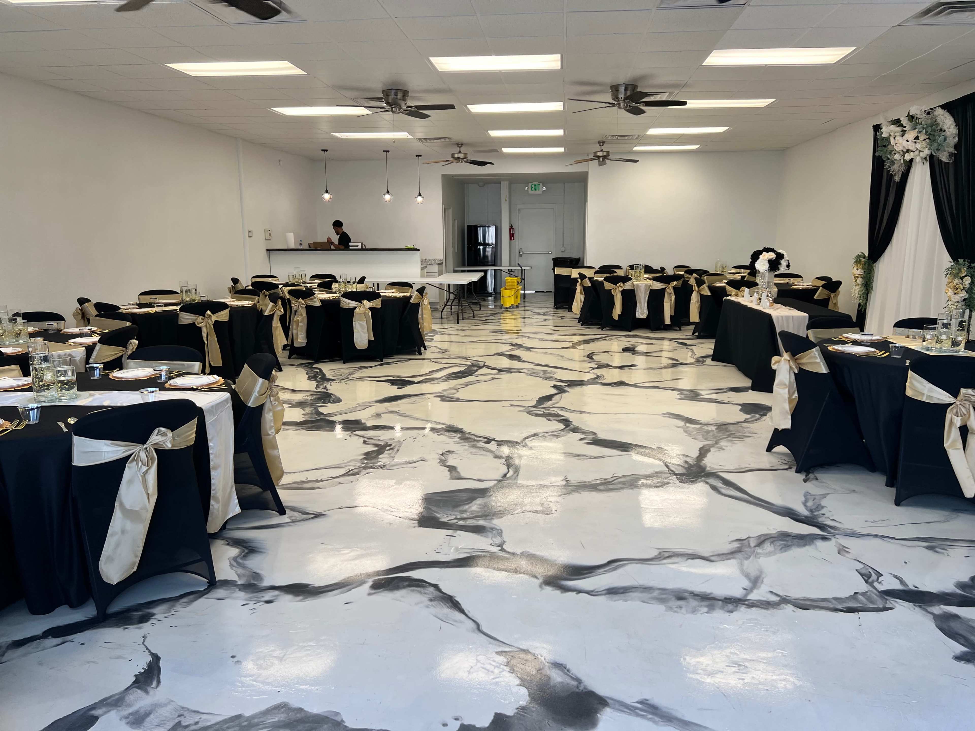 The room features neatly arranged round tables covered with black tablecloths and golden sashes, set against a marbled floor and minimalist decor.