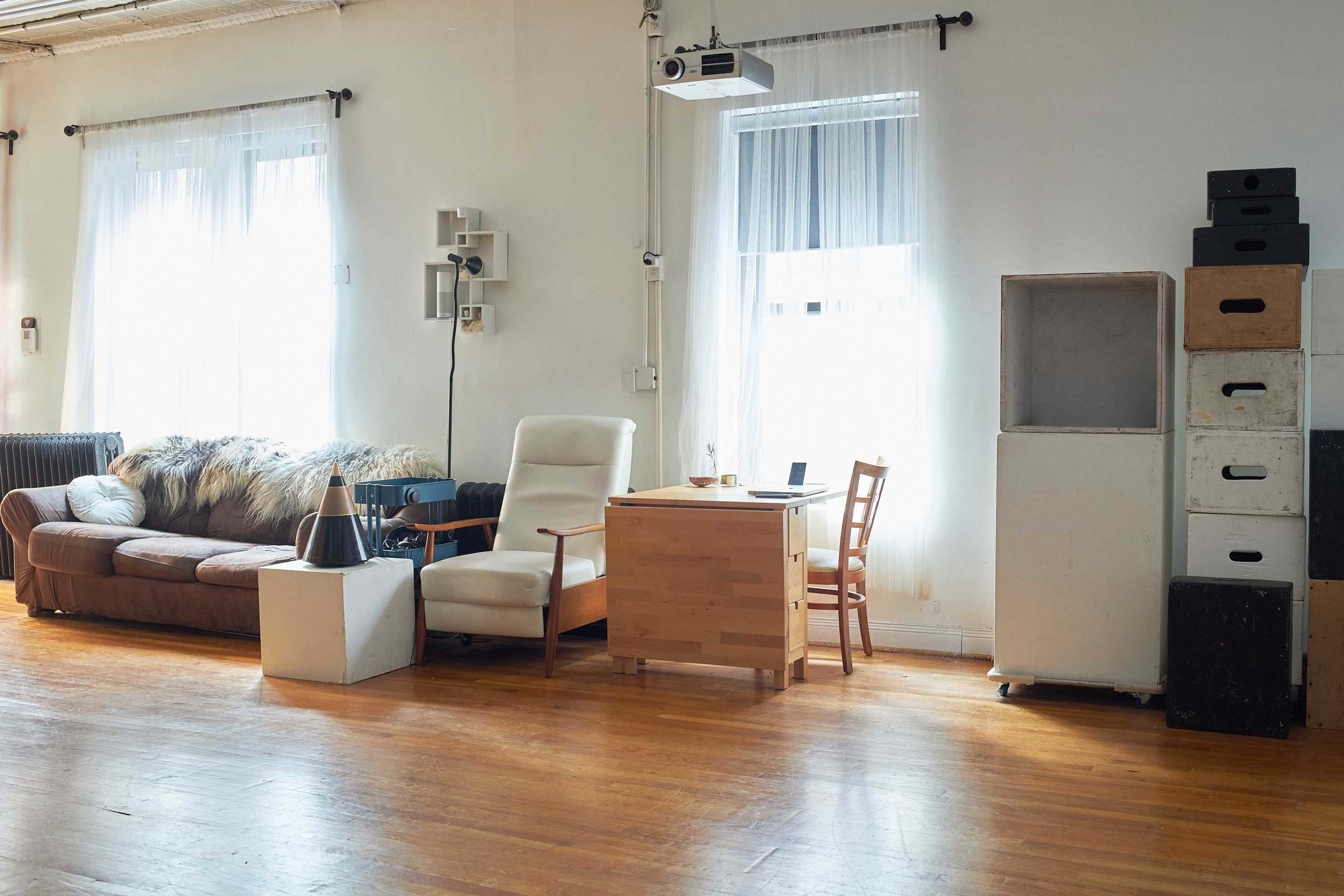 The image shows a spacious room with large windows, featuring a brown couch, a white armchair, a wooden desk with a chair, and storage boxes against the wall.