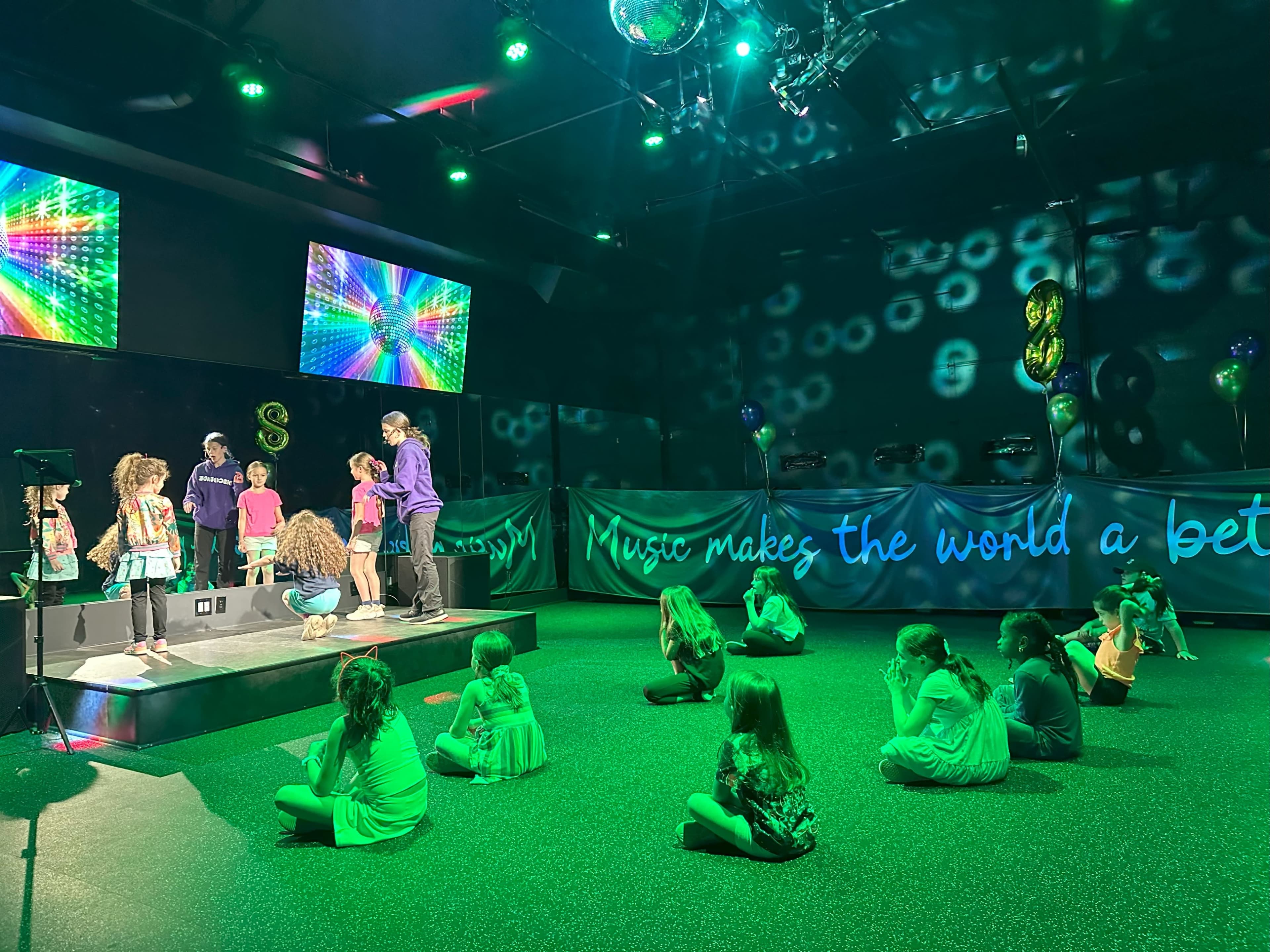 A group of children performs on stage with colorful lights and screens, while others sit on the floor watching.