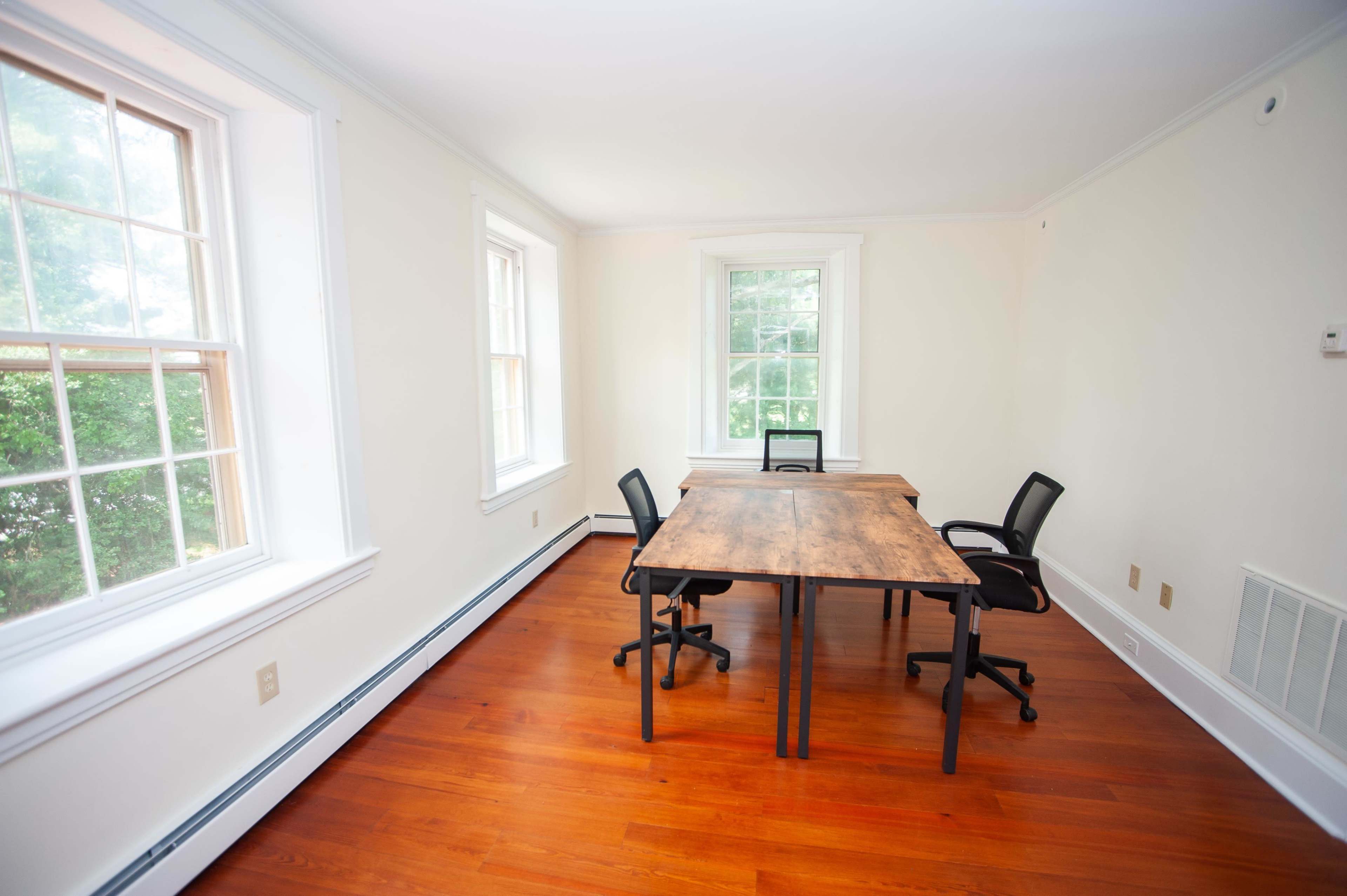 A bright, empty room with two large windows and a wooden table surrounded by four black chairs.