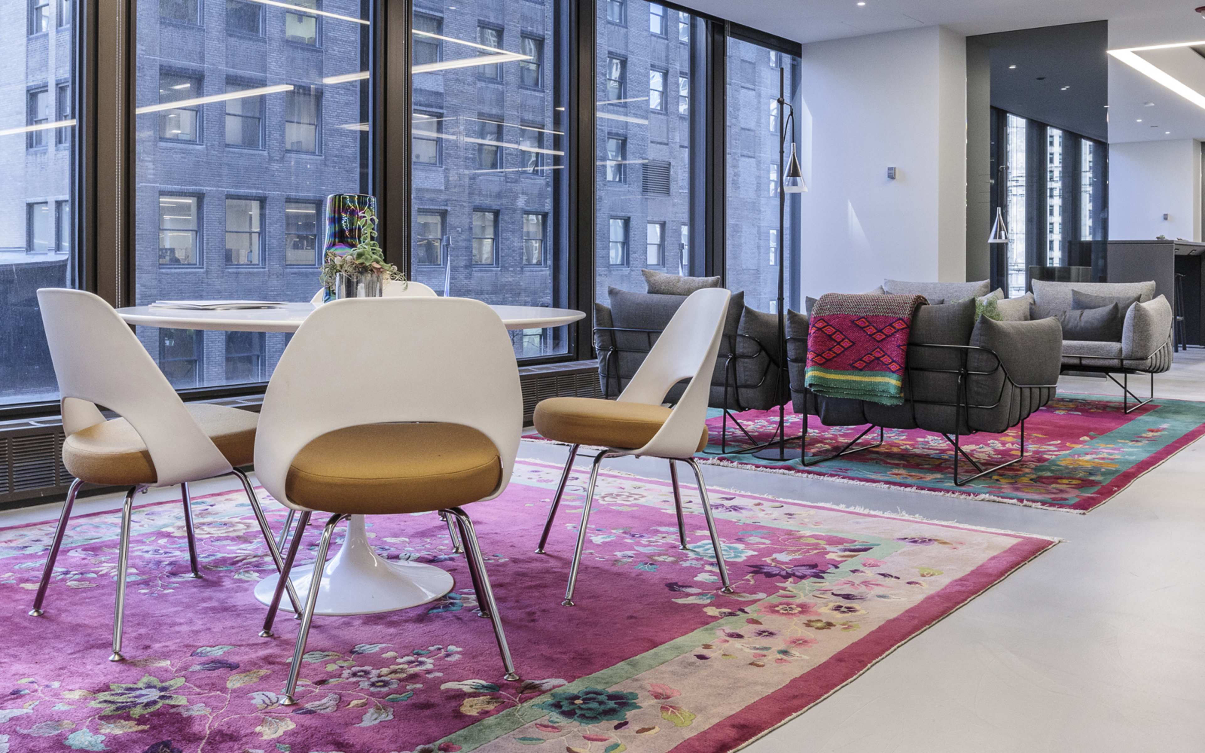 A modern office space features a round dining table with white chairs on a vibrant rug, alongside a seating area with gray sofas and large windows that provide city views.