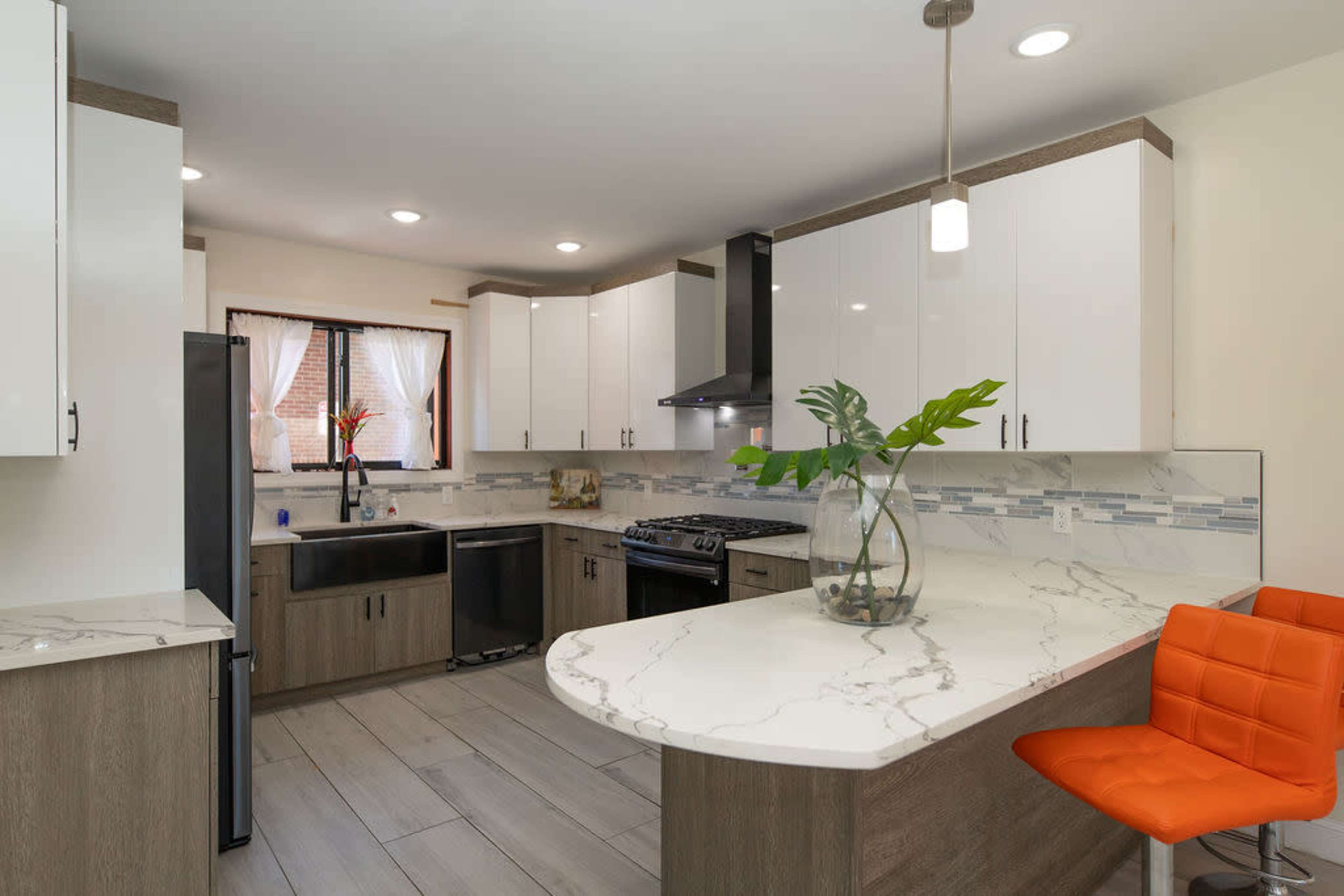 A modern kitchen with white cabinets, a gray countertop, and an orange bar stool at a curved island.