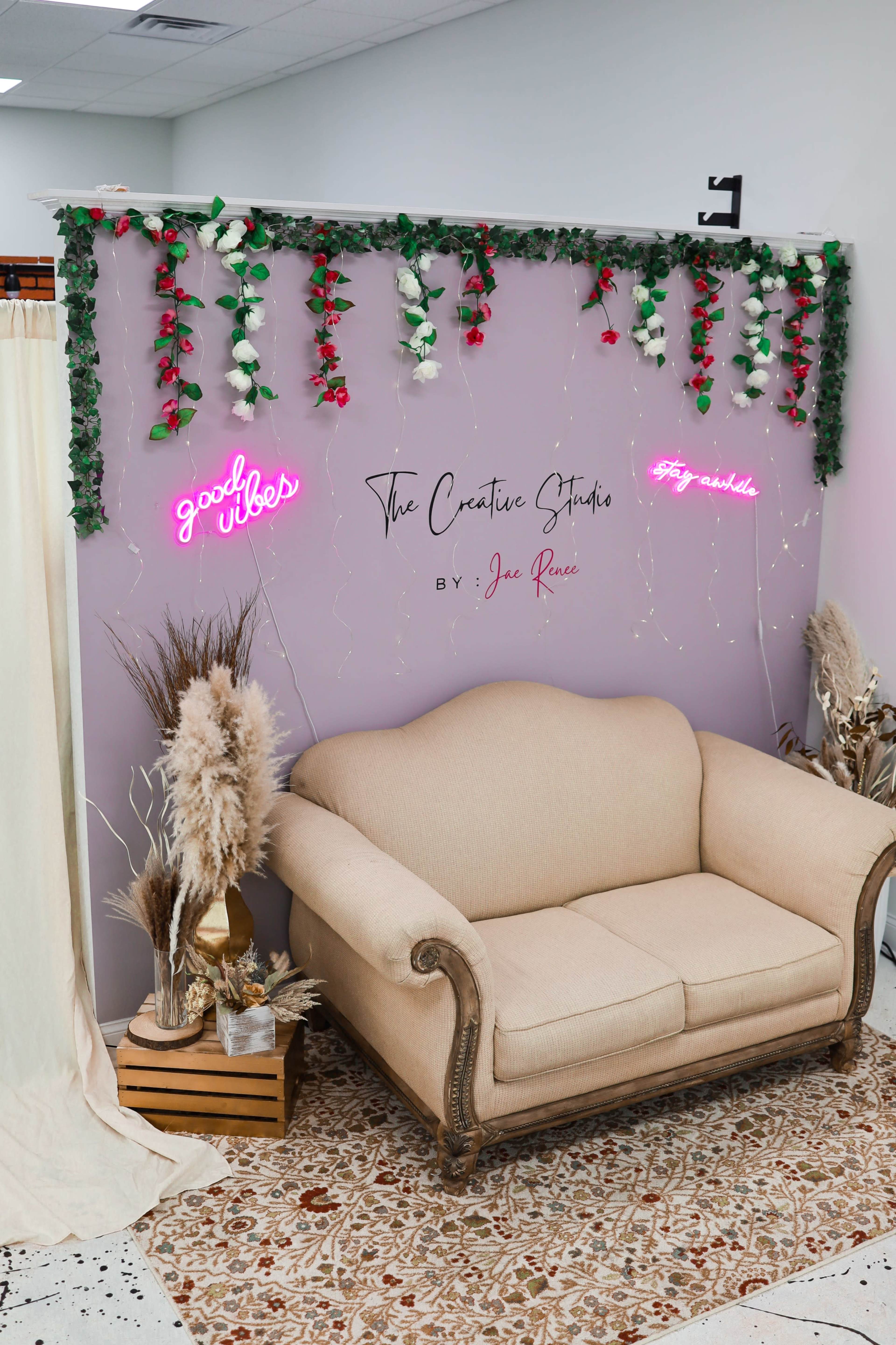 The image features a softly-lit interior with a beige couch against a pink wall adorned with green leaves and white roses, along with neon signs that read "good vibes" and "The Creative Studio."