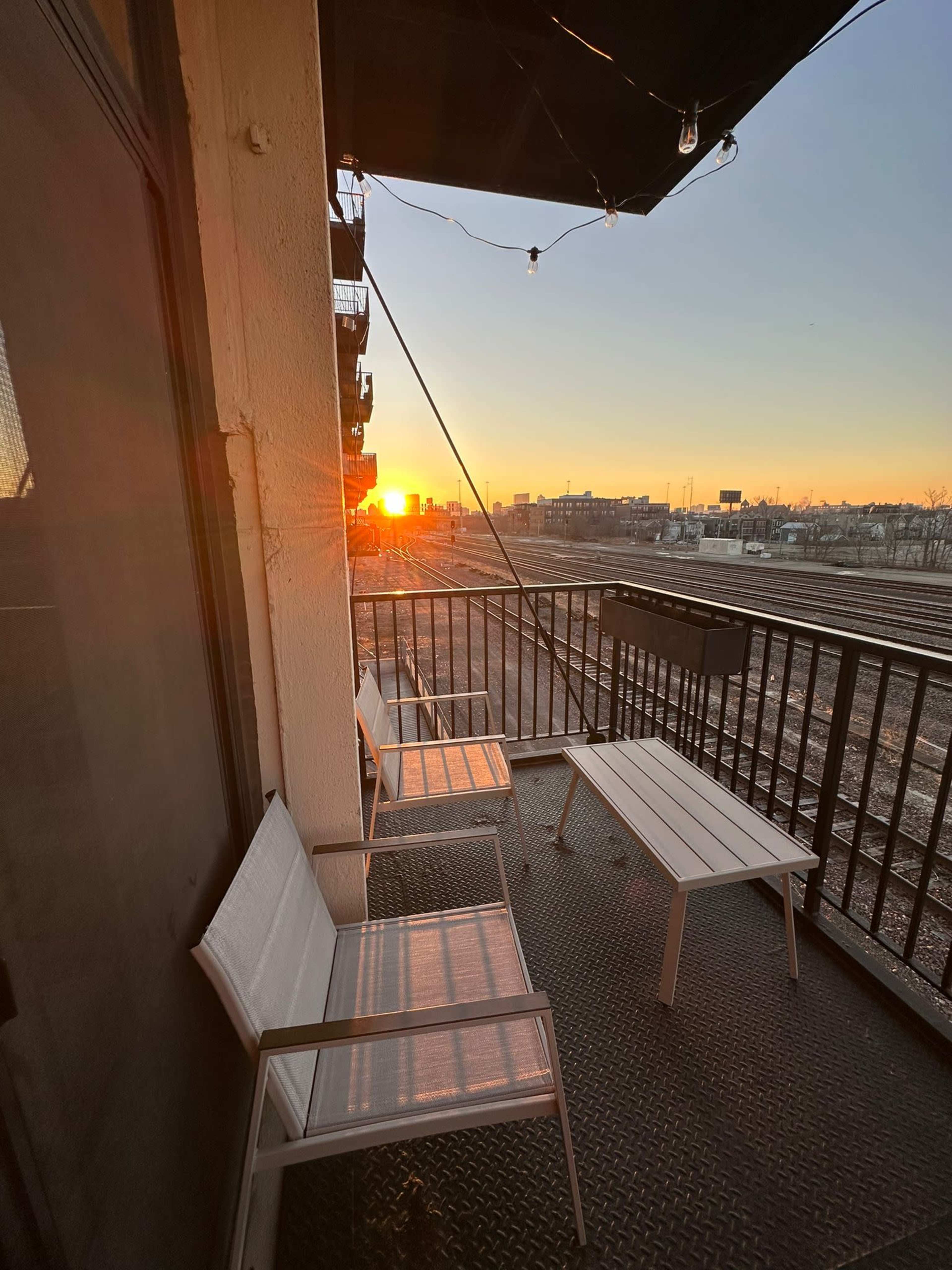 A balcony with two white chairs and a white table overlooks a sunset against a cityscape.